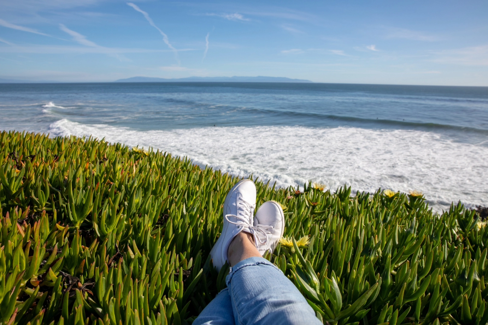 Person wearing jeans and white sneakers sits by the sea on a sunny day, enjoying the solitude and time alone by themselves.