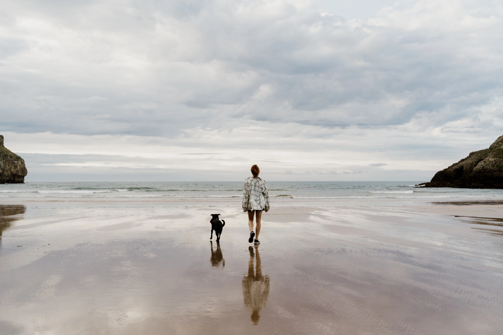 Woman and dog walk along a quiet beach during the day 