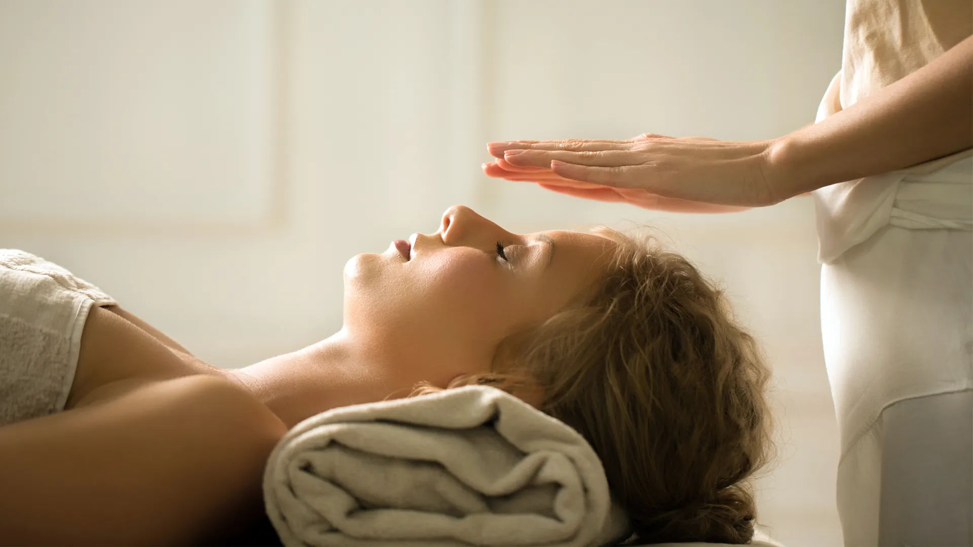 A woman lying on a table with her head resting on a white rolled up towel receives reiki treatment. 