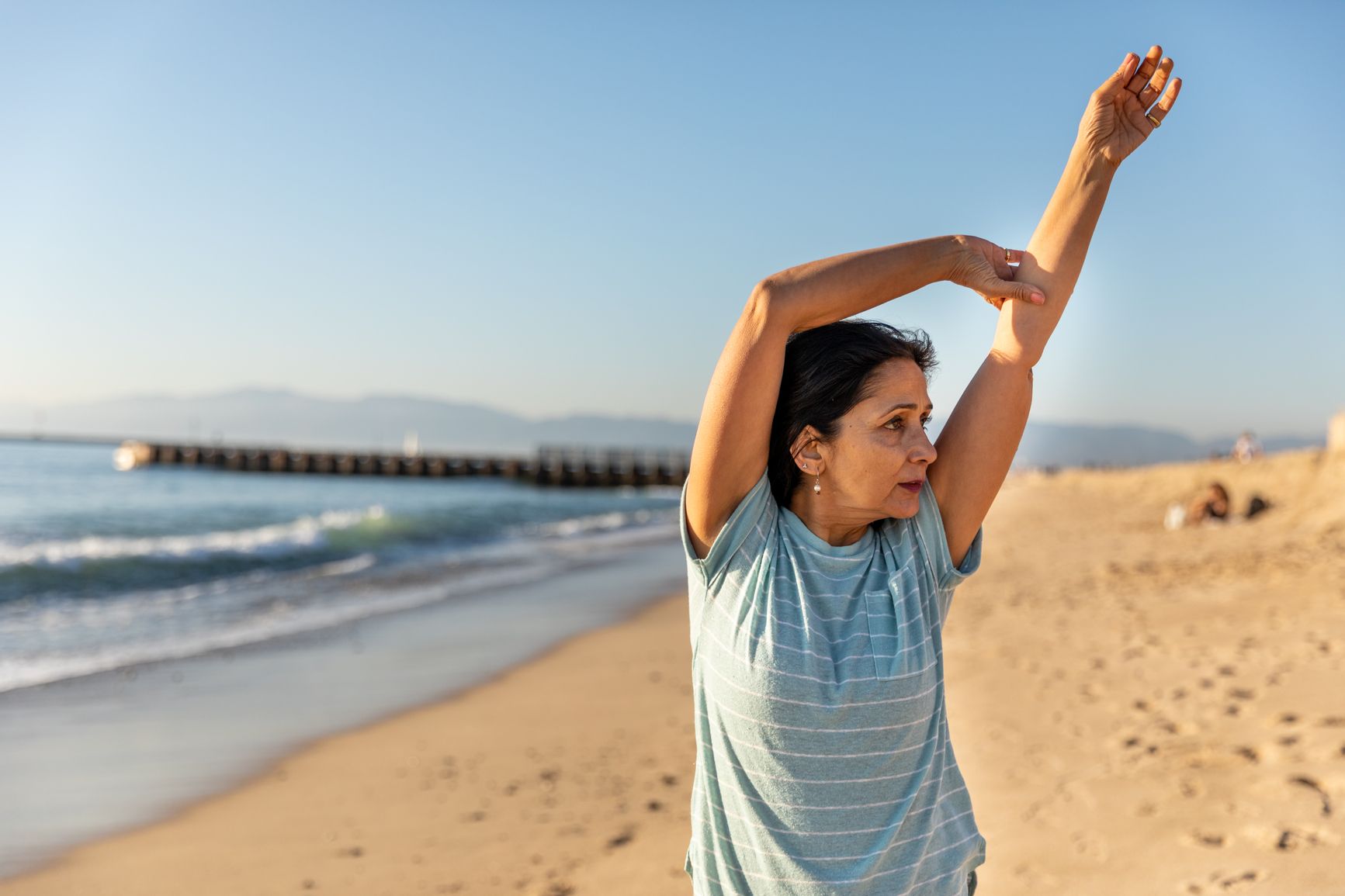 Woman on the beach wearing a striped t-shirt gently stretches her arms overhead 