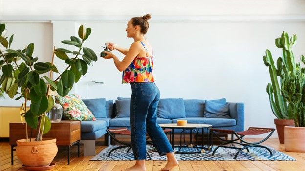 White woman in colorful tank top and jeans sprays water on tall plant in her living room.