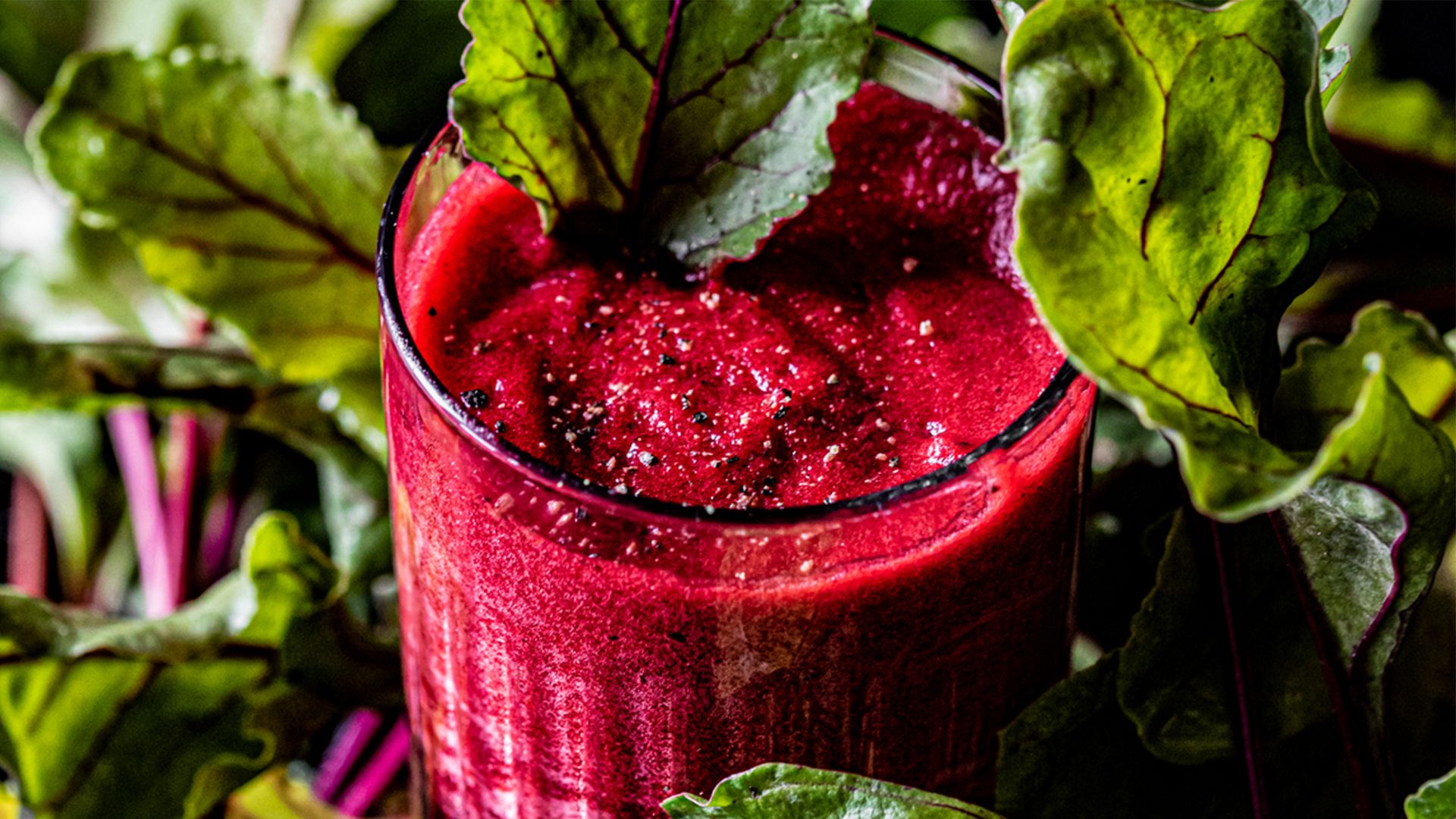 A close-up photograph of a glass of bright red beet juice with spinach garnishes set a background of leafy greens.