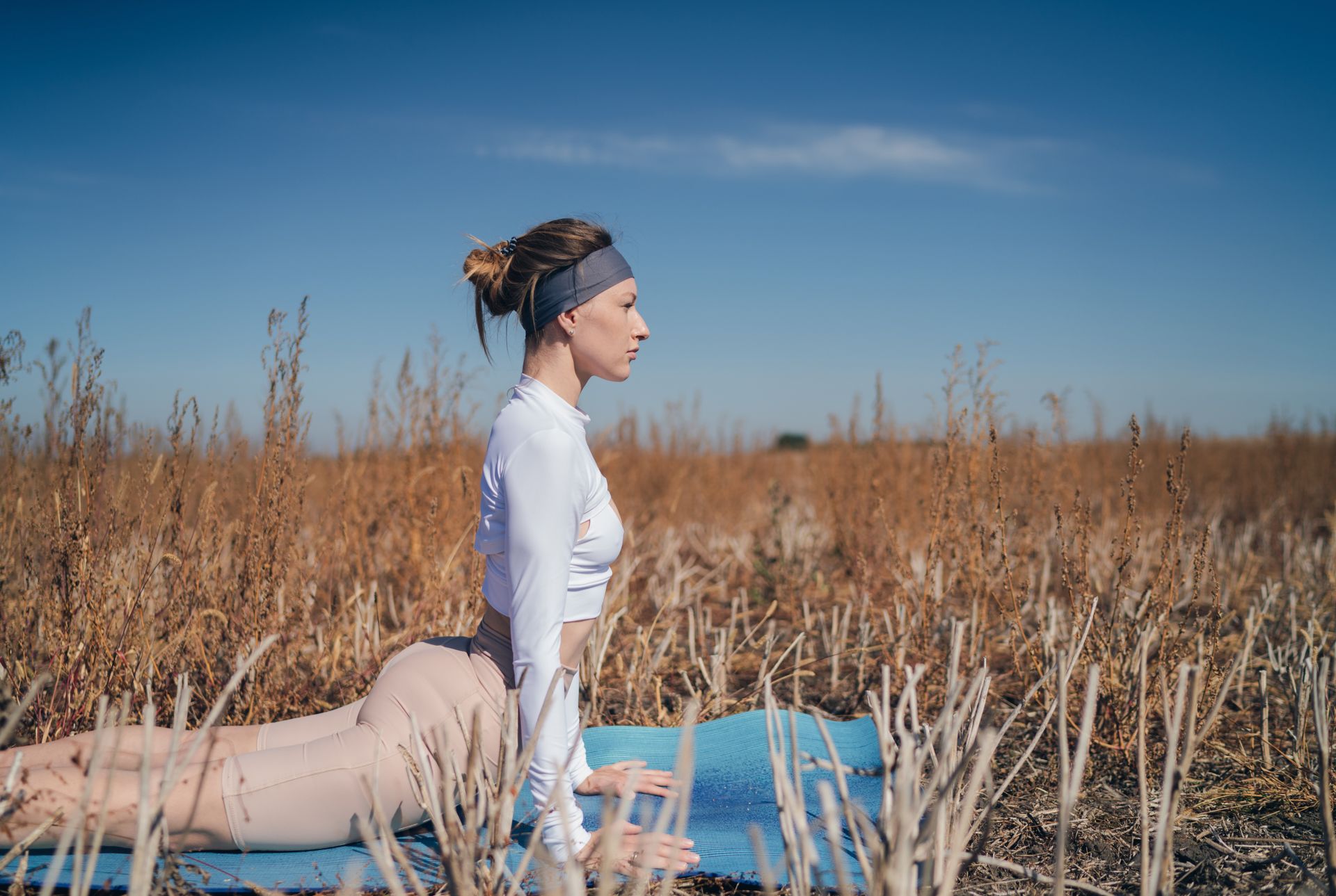 Woman in white long-sleeve shirt does an upward dog pose on a mat in an outdoor field 