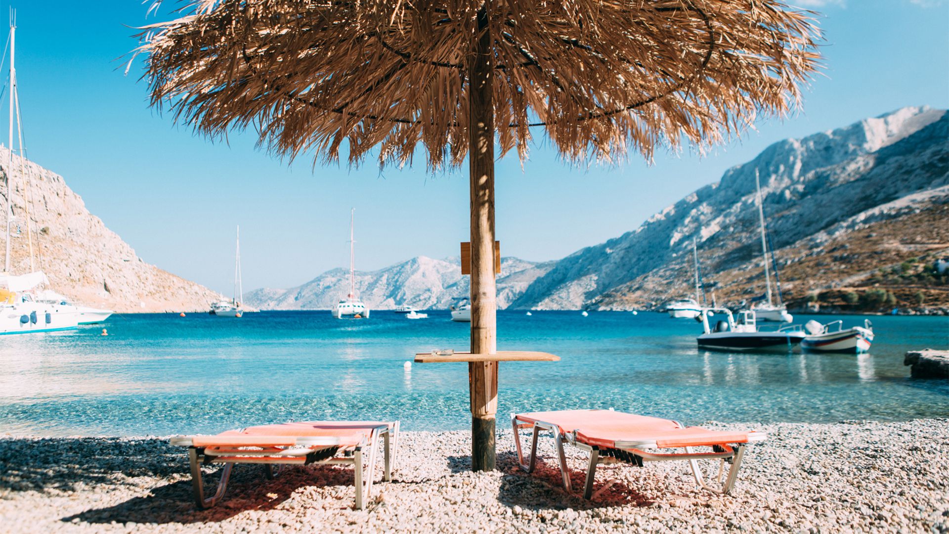 Photograph of a beautiful beach, with sunny, blue skies, boats in the water, and mountains in the background. In the foreground, two pink beach lounge chairs lay open underneath a large straw umbrella.