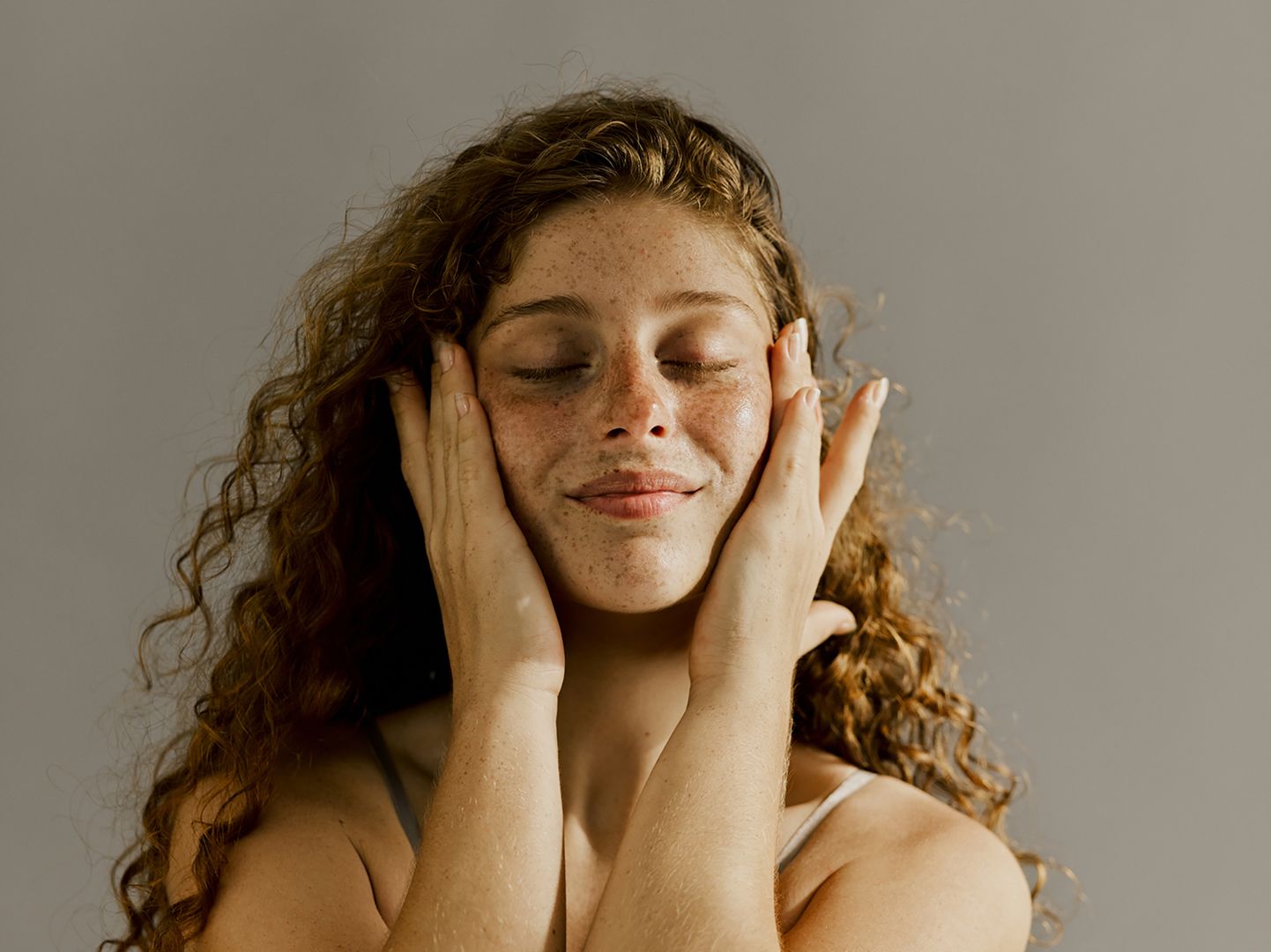 Young white woman with long curly hair looking relaxed massages her face with her fingers.