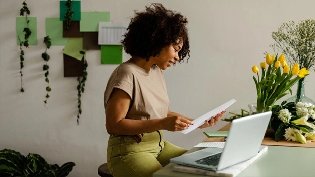 Black woman wearing green pants and a tan top. Sitting at her desk holding a pen and a notepad.