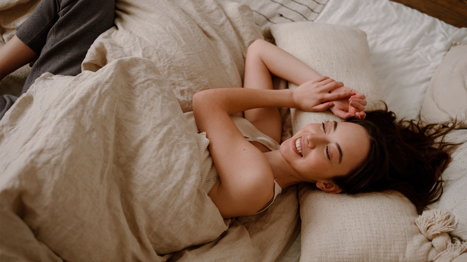 Young woman smiles while lying in bed under rumpled beige sheets.