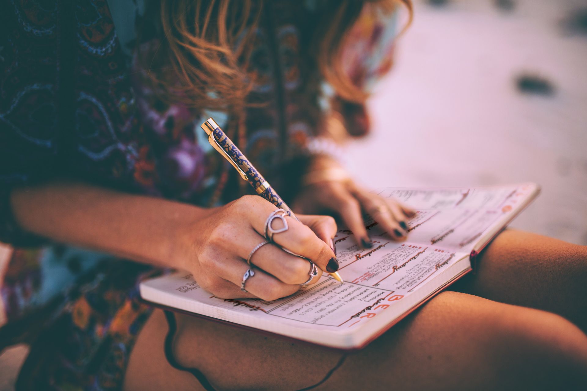 A woman wearing a floral dress writes in a paper journal that she holds on her lap.