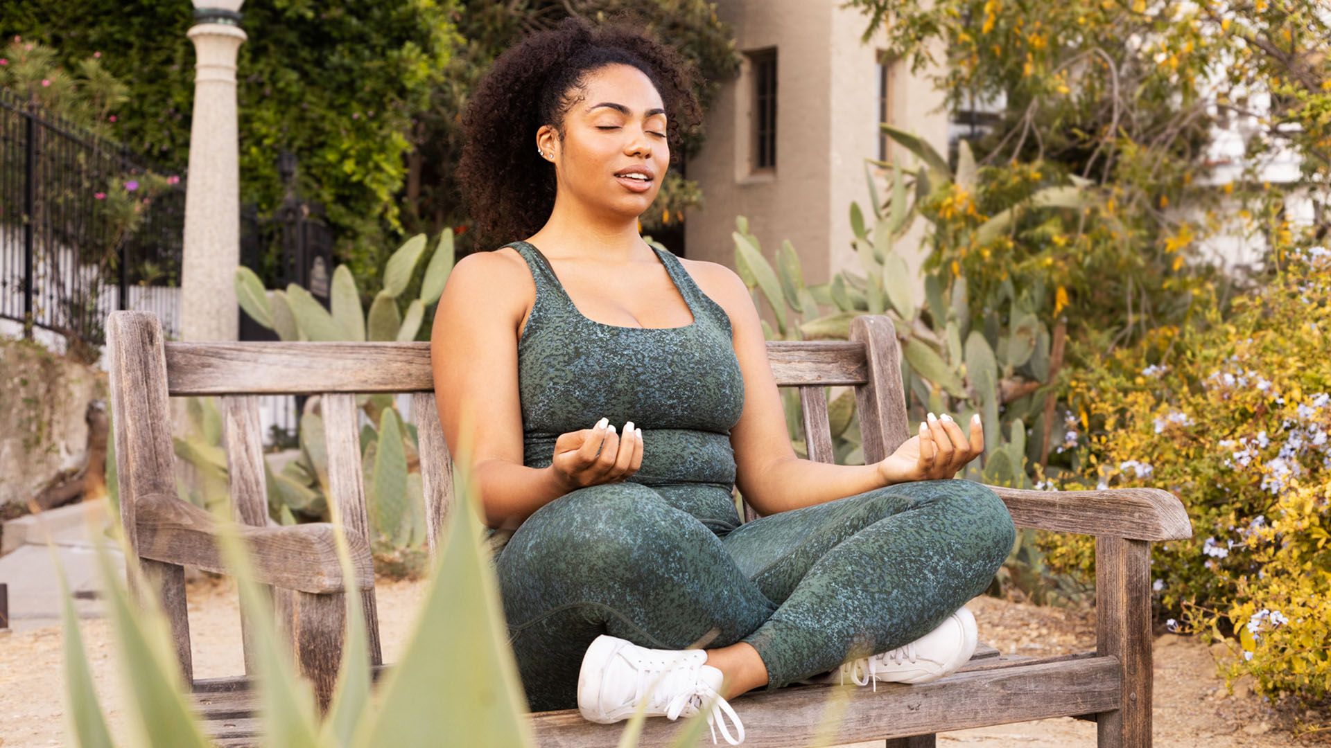 Woman in exercise clothing and white sneakers sits cross-legged with her eyes closed and meditates peacefully on bench in outdoor cactus garden