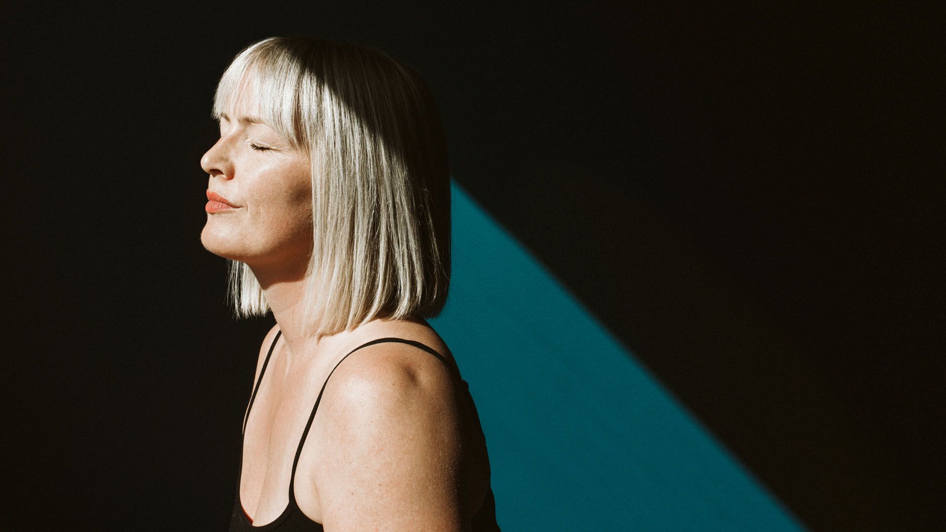 Woman with medium-length straight blonde hair stands near a blue wall, her face lit by the sun and her eyes closed, looking peaceful and content. 