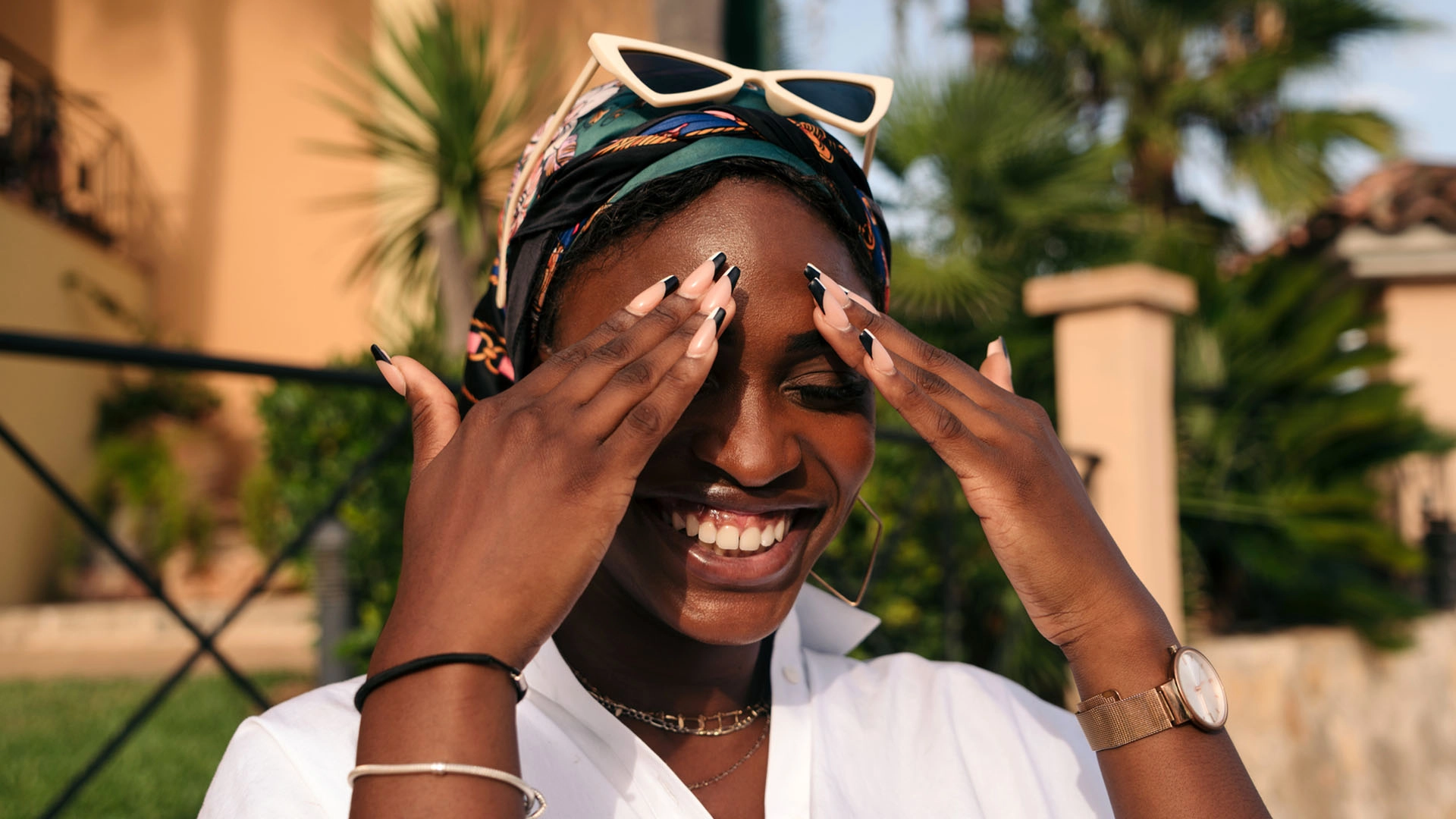 A Black woman standing outdoors in the sun holds her hands to forehead and smiles broadly, looking happy and content.