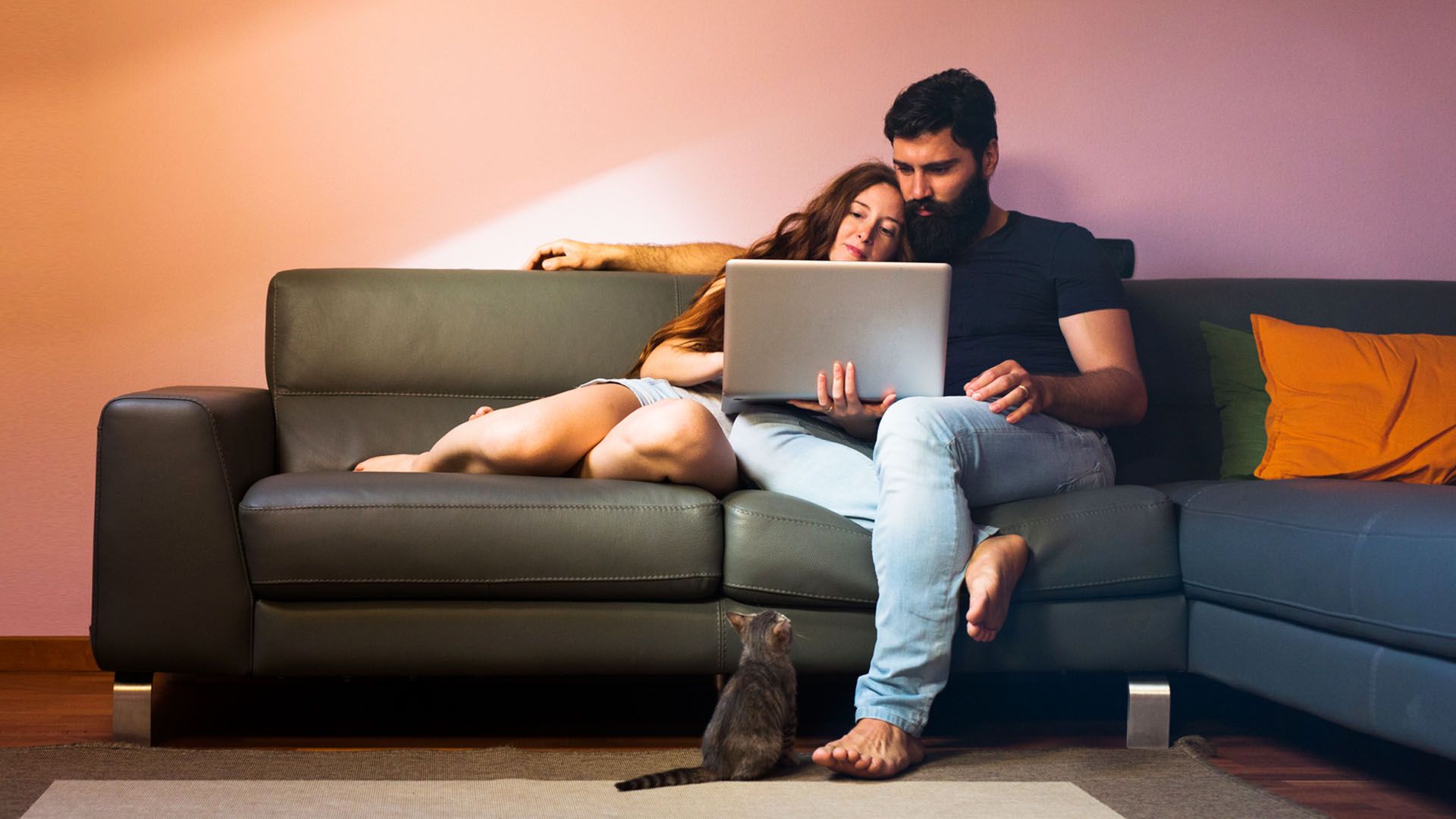 Couple sit together on a couch snuggling and looking at a computer while a small cat sitting on the floor looks up at them.