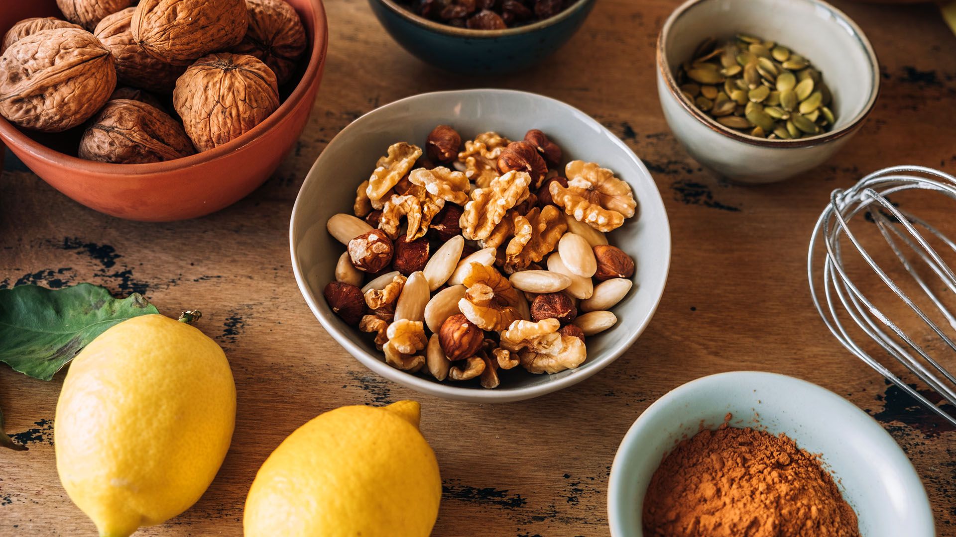 Bowls of snacks such as walnuts, almonds, and pumpkin seeds on a wooden table rest next to a whisk and lemons.