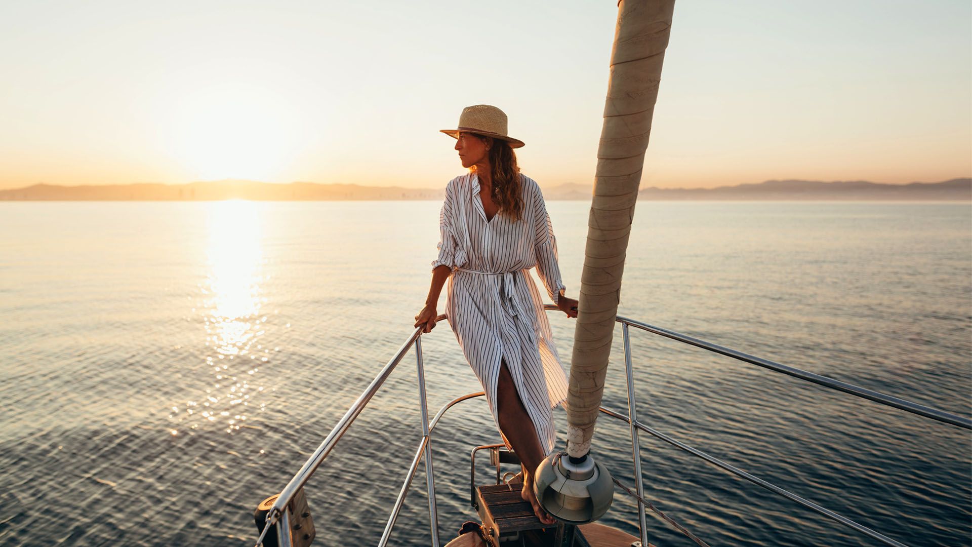 Woman wearing striped dress and straw hat stands on the deck of a sailbox enjoying the sunset.