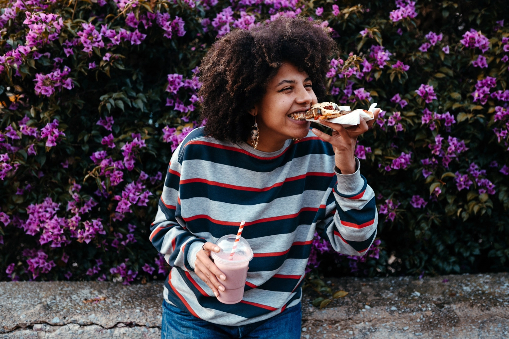 A woman standing in front of a purple flower bush, smiling happily as she eats pizza and drinks a pink milkshake