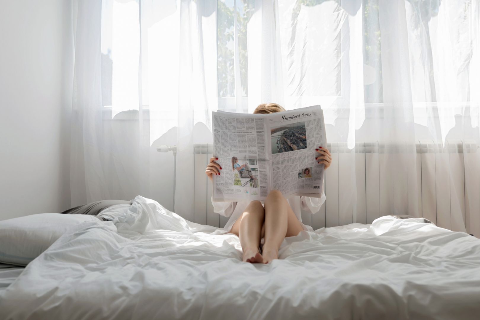 Person sits on top of a white comforter in bed reading a daily newspaper.