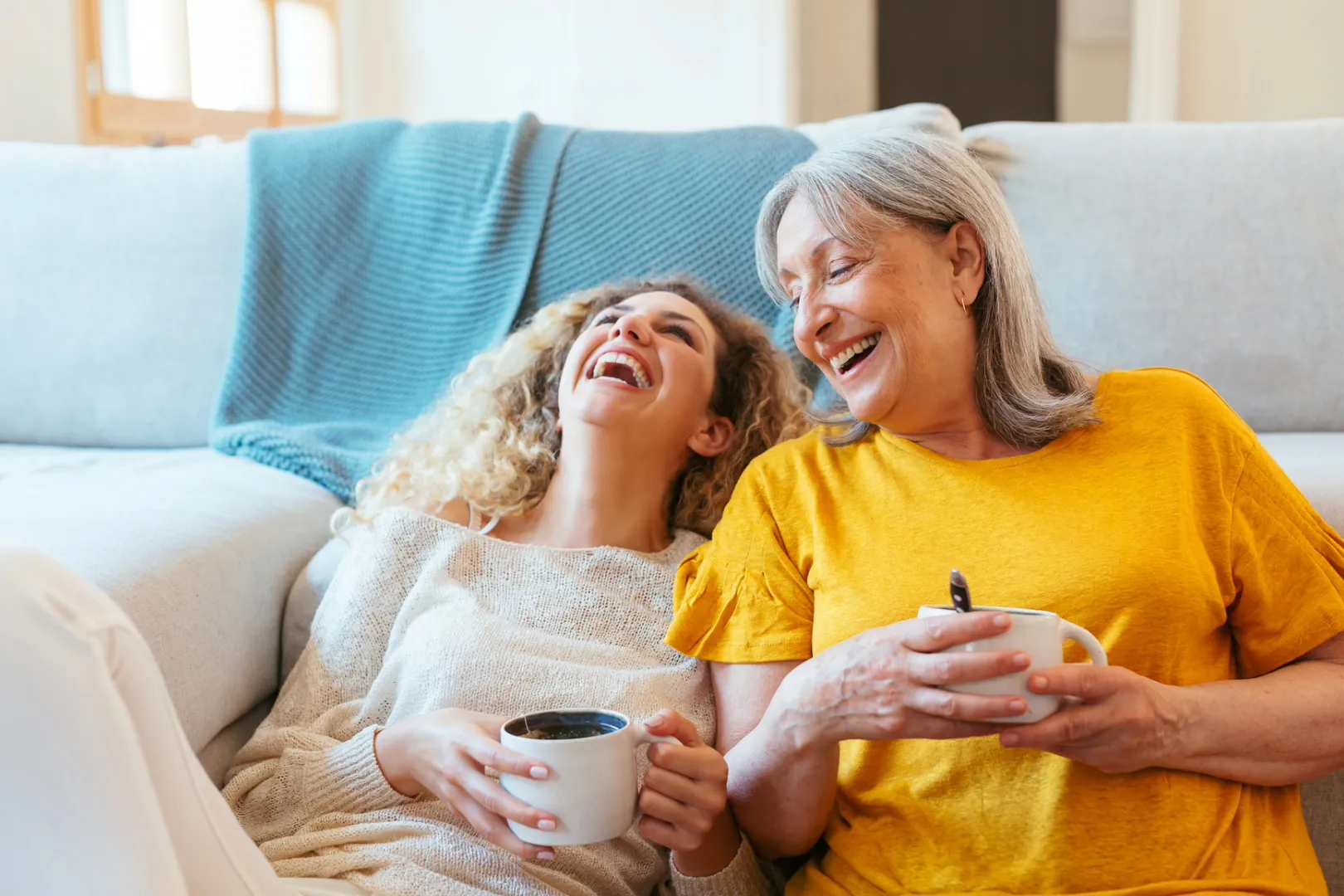 A mother and daughter sit leaning against a couch in a sunny living room, laughing together with their heads closer together.