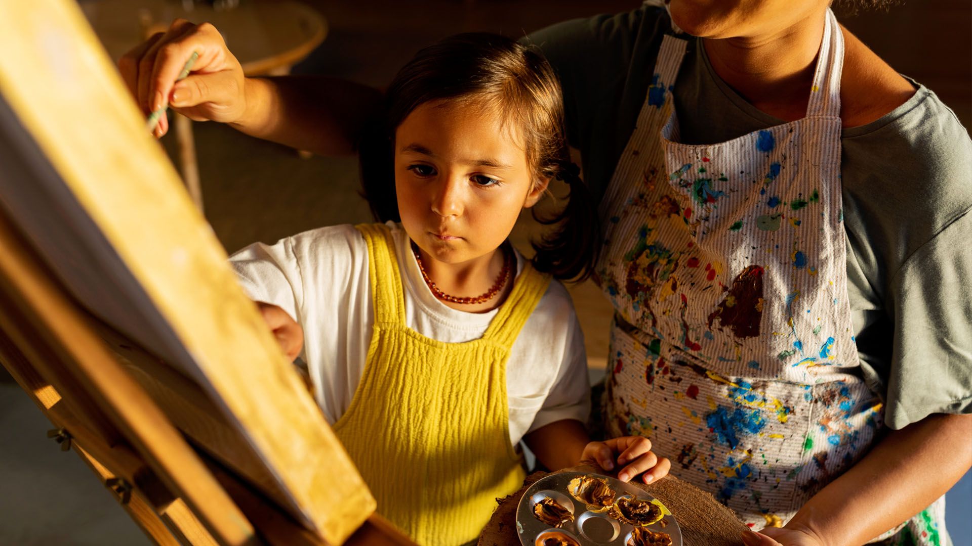 Mother and young daughter stand together in front of an easel painting a picture.