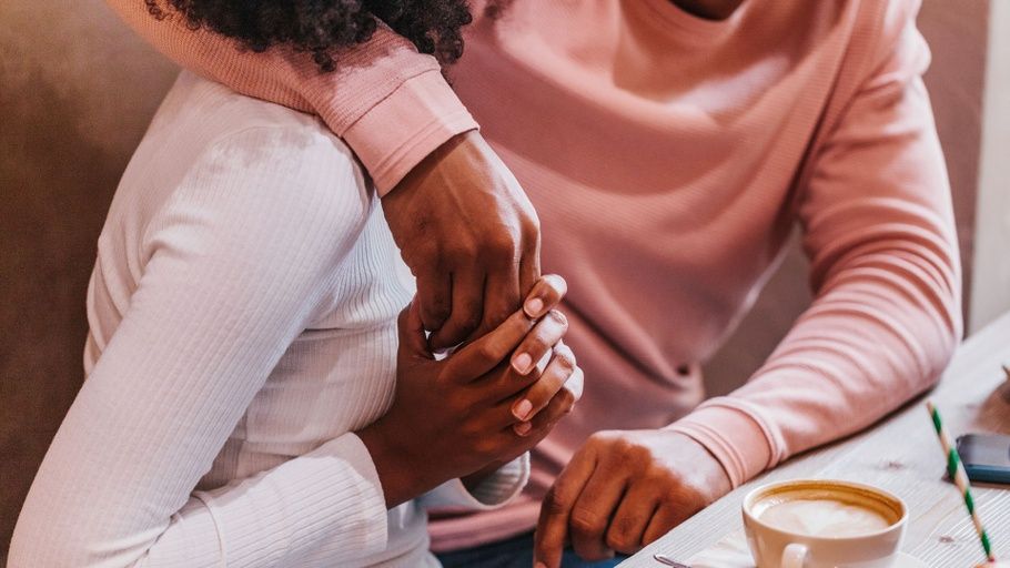 A couple, shown from the shoulders down, and both wearing tops in shades of pink, sit together holding hands in a cafe.