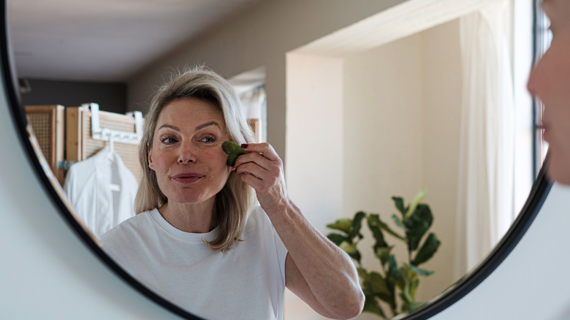 Woman stands in front of a mirror holding a gua sha tool as part of her facial fitness routine.