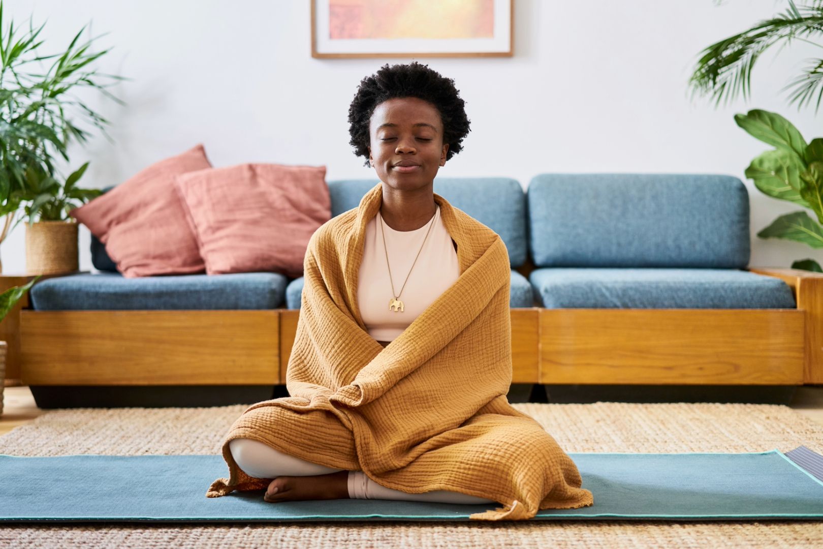 Woman sits crossed-legged on a living room floor, wrapped in a blanket, eyes closed as she calmly repeats peaceful affirmations to reduce stress