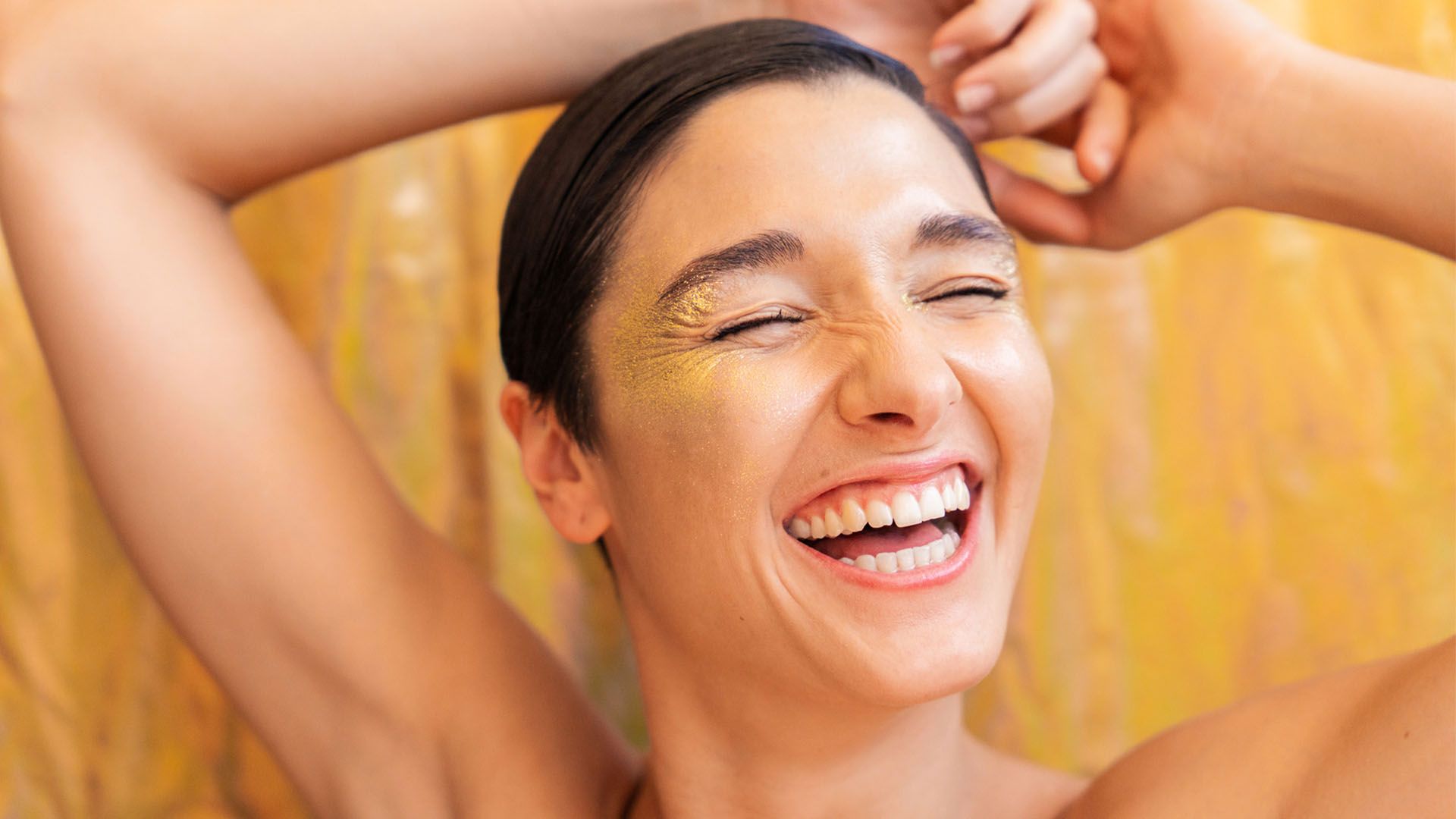 A close up of a woman with short brown hair as she smiles and laughs, eyes closed, while her arms are raised above her head.