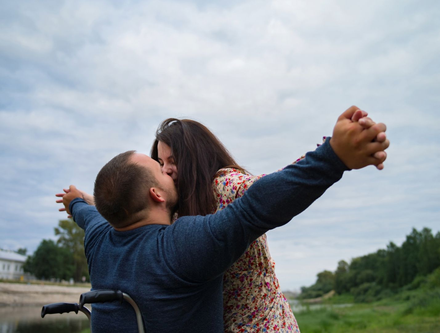 A man in a wheelchair and a woman smiling with her arms outstretched kiss and embrace with the blue sky and green trees behind them.