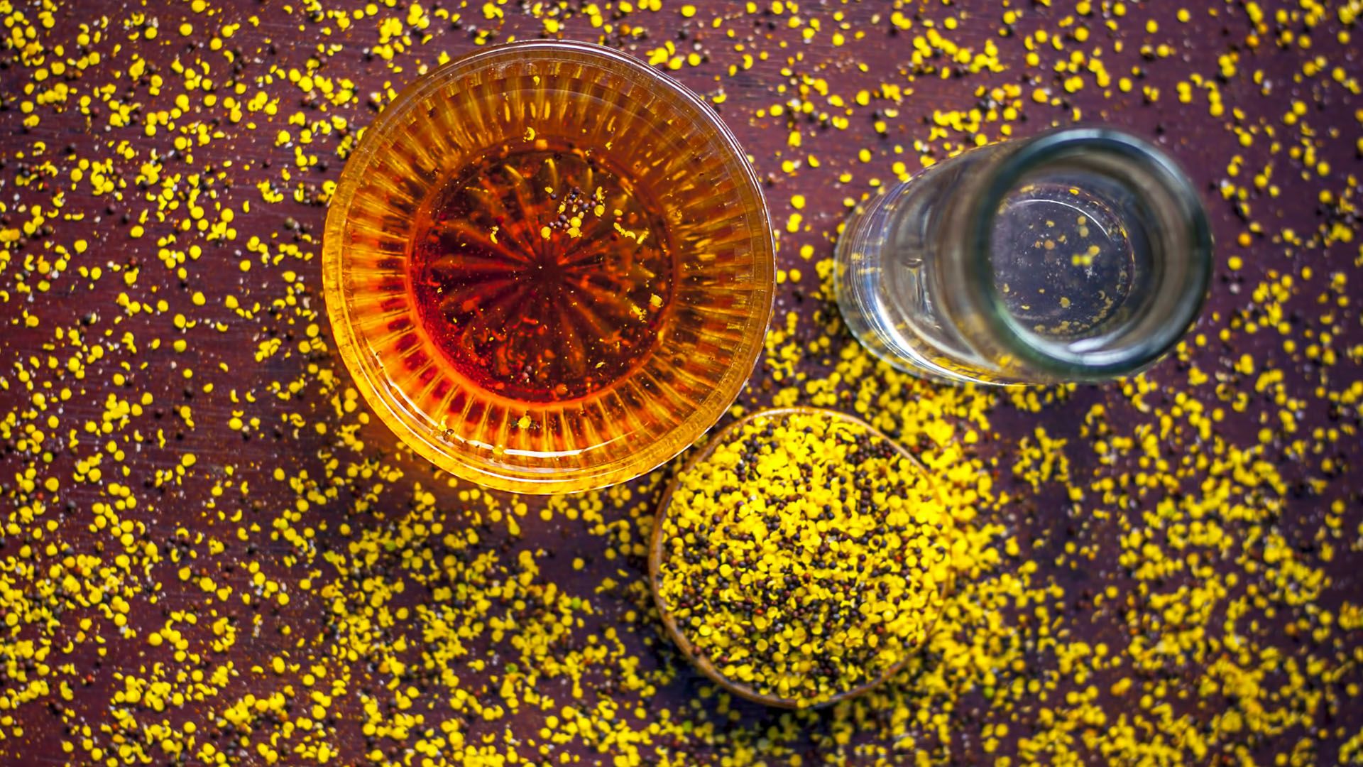 Two bowls, one with crushed brown mustard seeds and the other with honey are photographed alongside a glass of water.