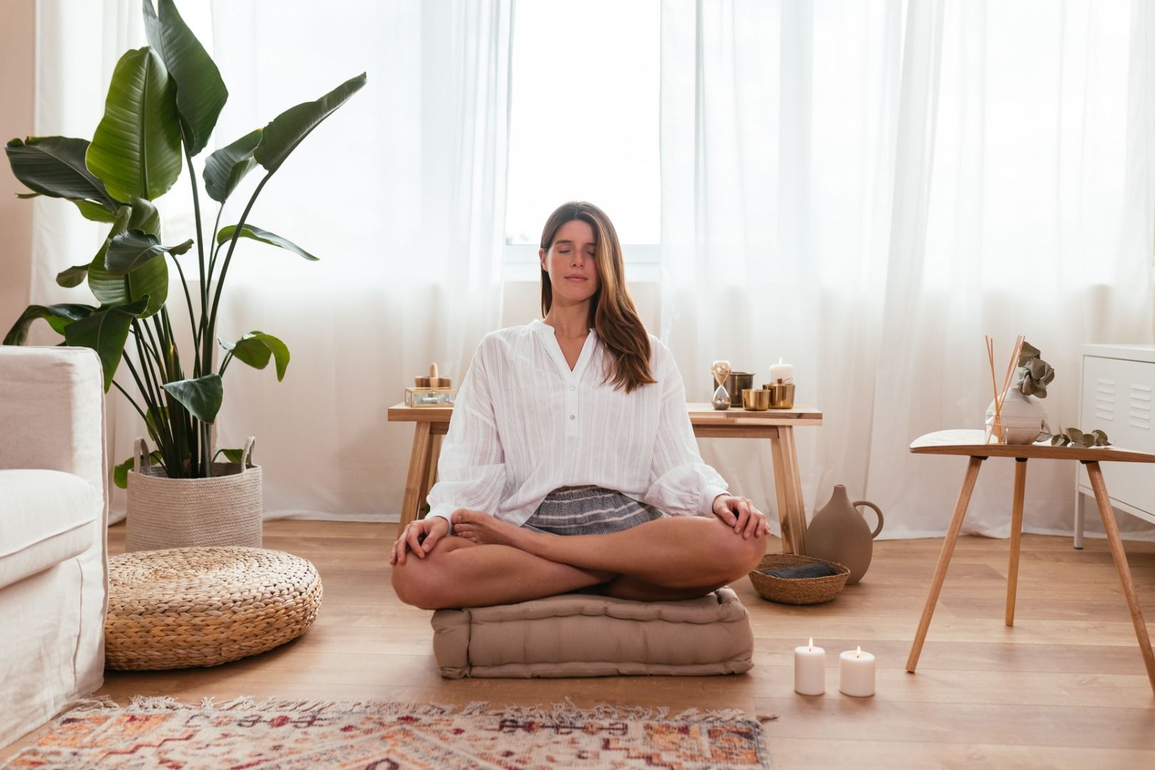 In a bright, sunny living room, a woman sits on a pillow with her legs crossed and eyes closed, meditating to channel her spirit guide.