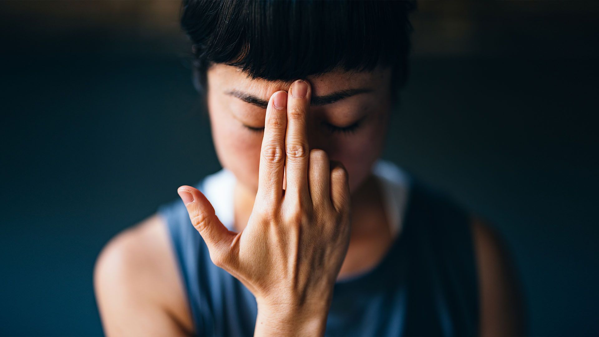 Close-up photo of a woman wearing a blue tank top using her index and middle finger to tap on her forehead.