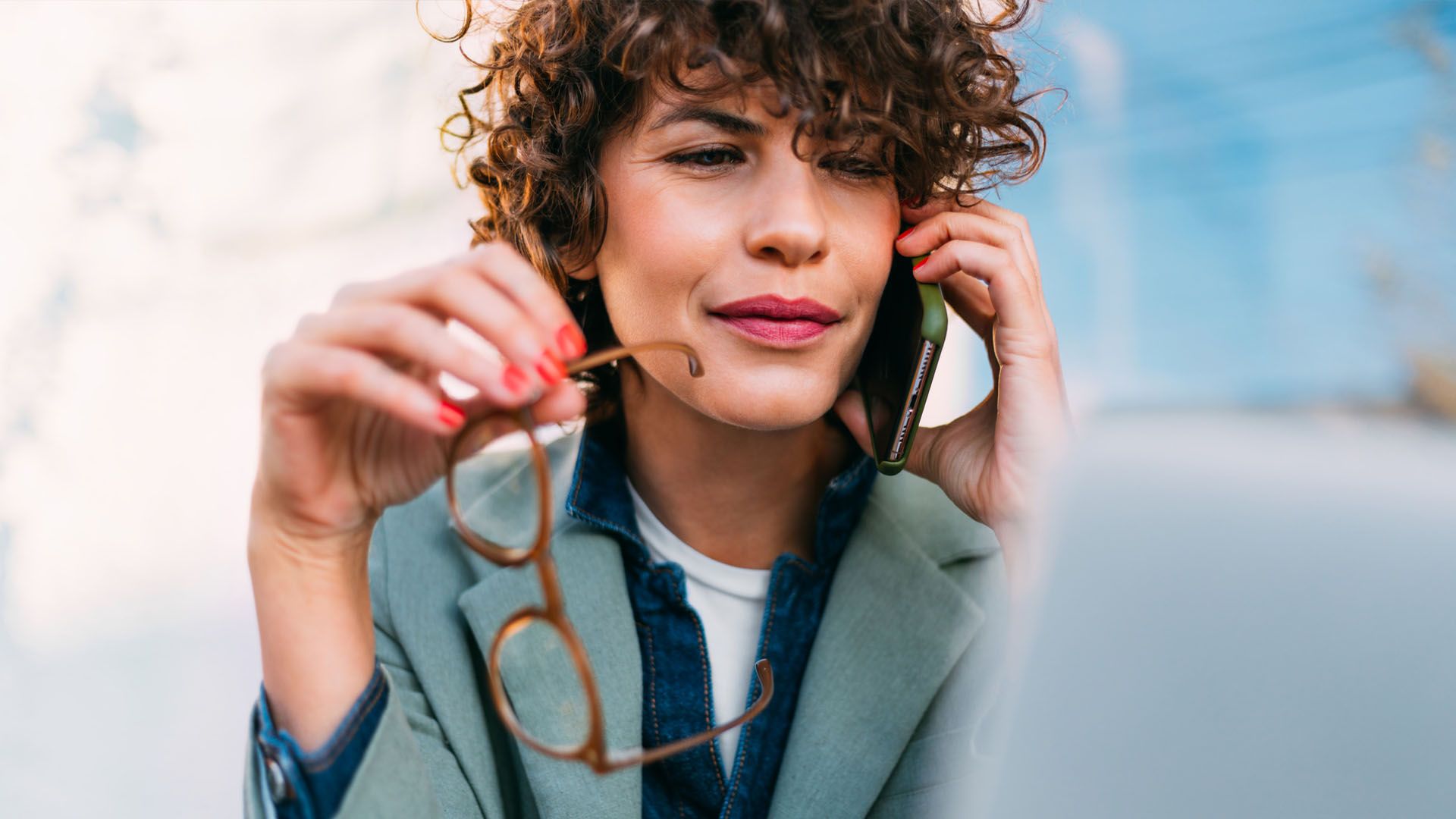 Woman wearing business casual clothing holds her cell phone in one hand while holding her eyeglasses in the other.