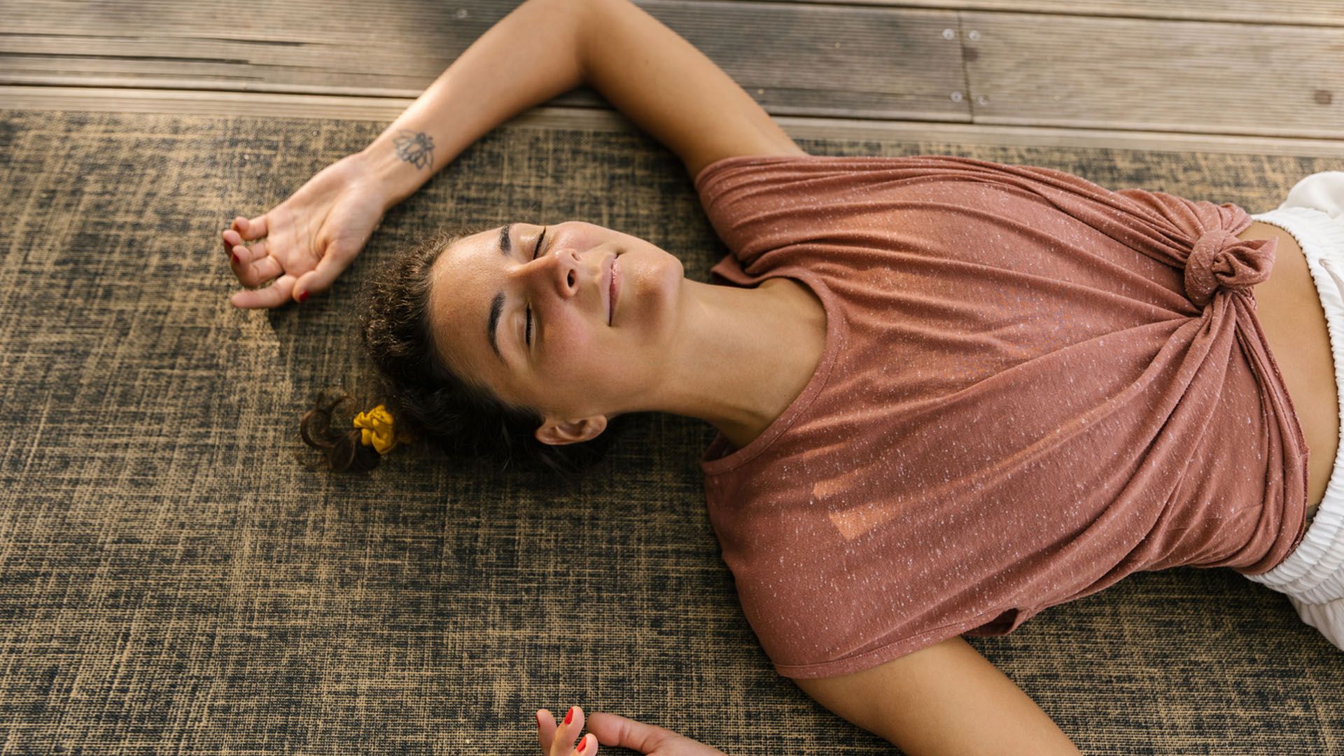 White woman with a tattoo on her wrist and wearing a pink t-shirt lays on the floor, with her eyes closed and a smile on her face, resting contentedly.