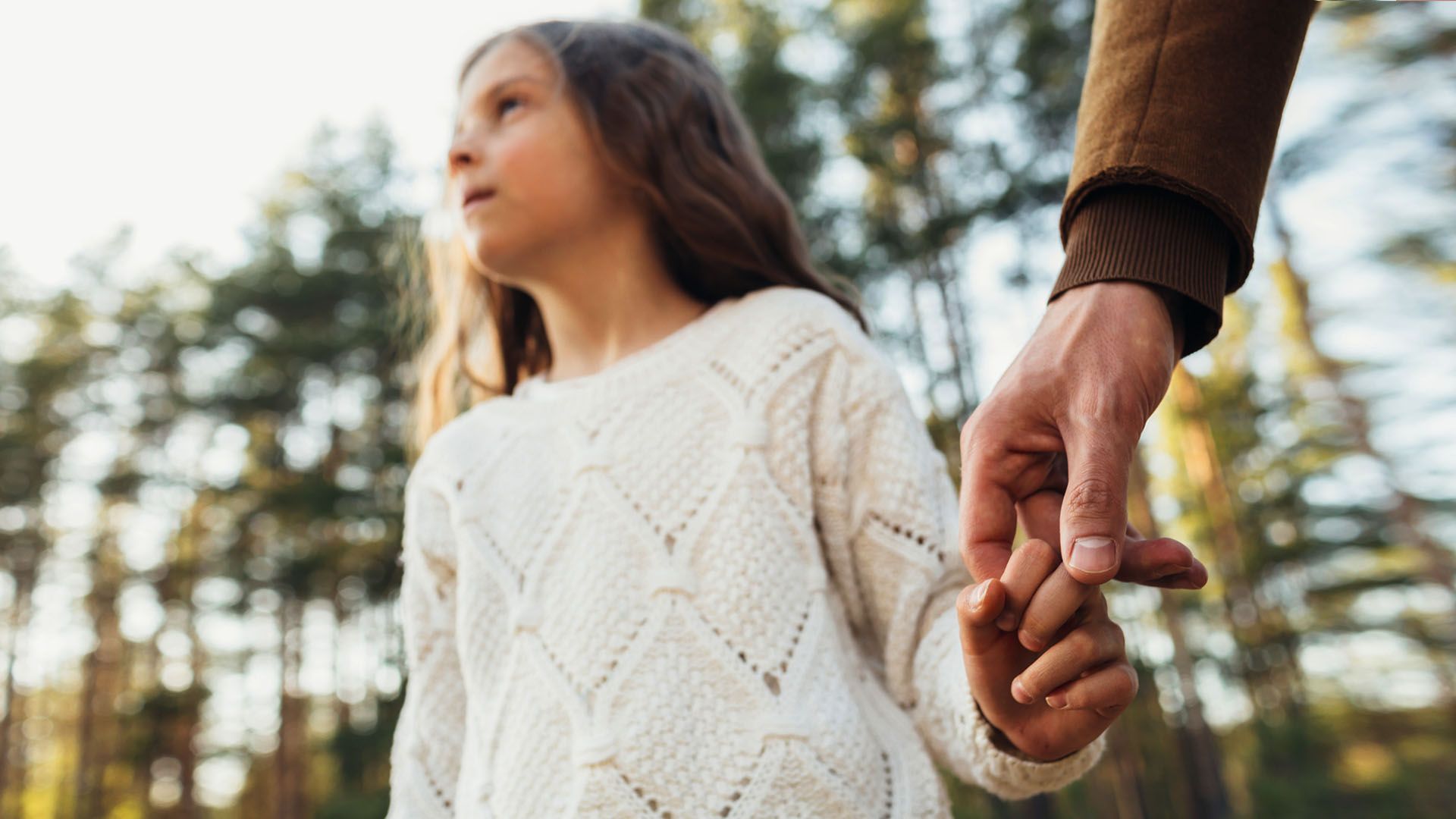 Little girl wearing a white sweater with a serious expression on her face holds her father’s hand, with a closeup on hands