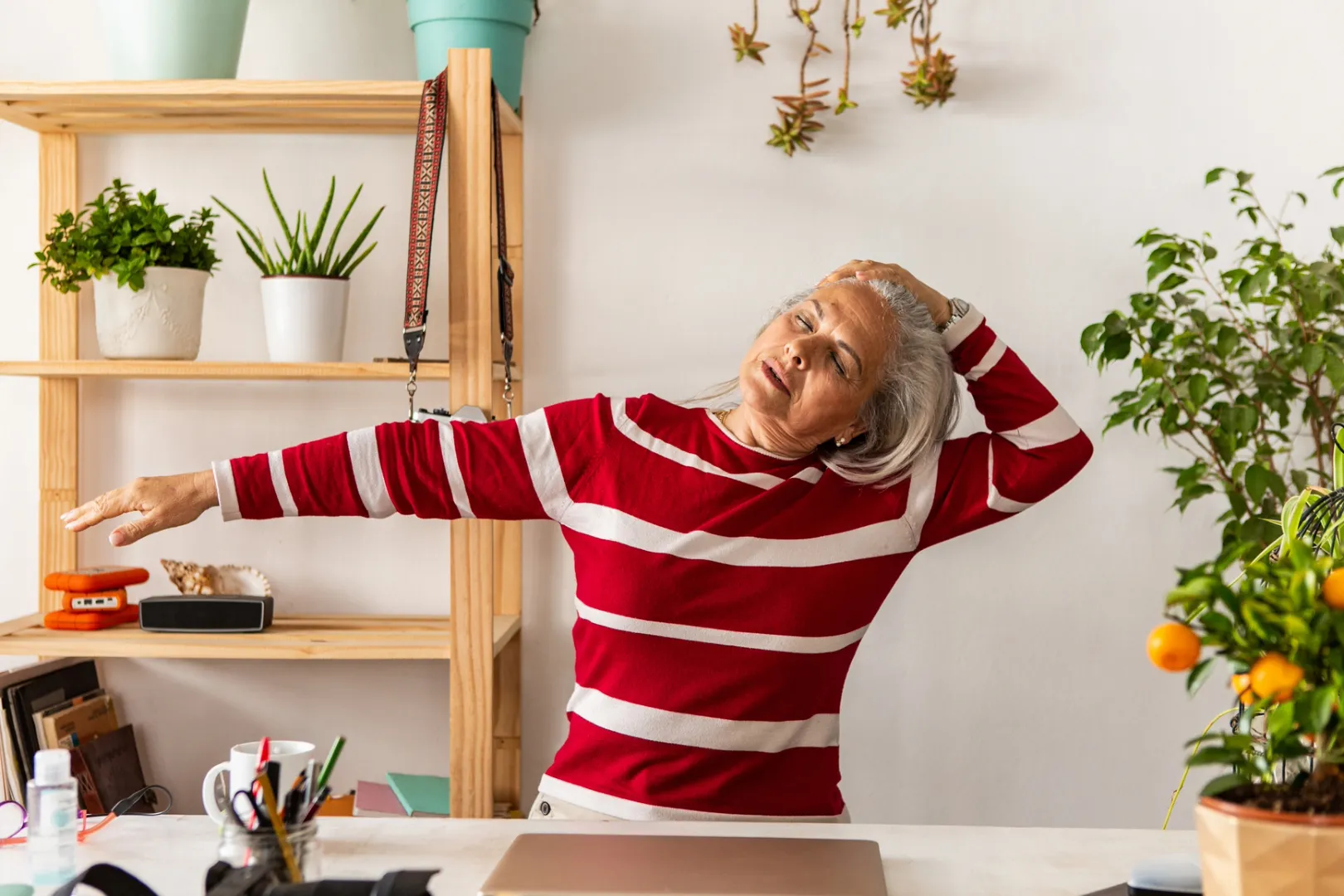 Senior woman wearing a red and white striped shirt closes her eyes and performs a gentle Feldenkrais Method stretch while sitting at her desk.