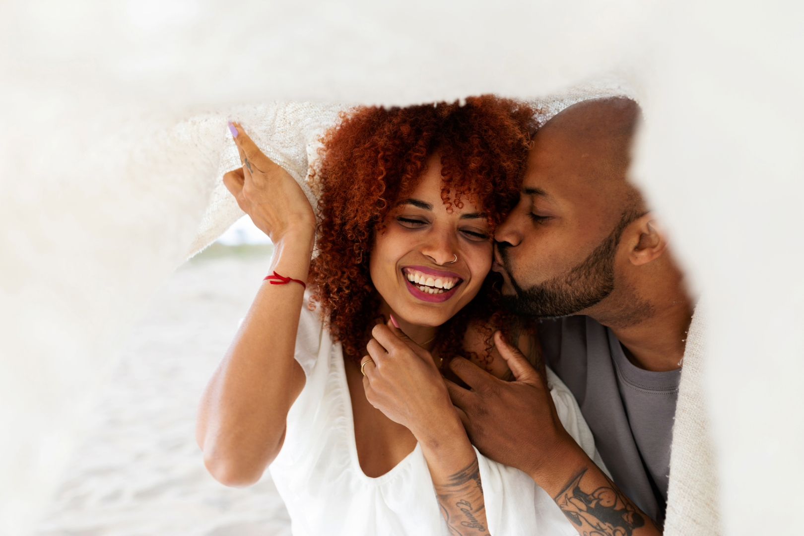 A happy Gen X couple laughs and kisses while snuggling in bed under a white sheet