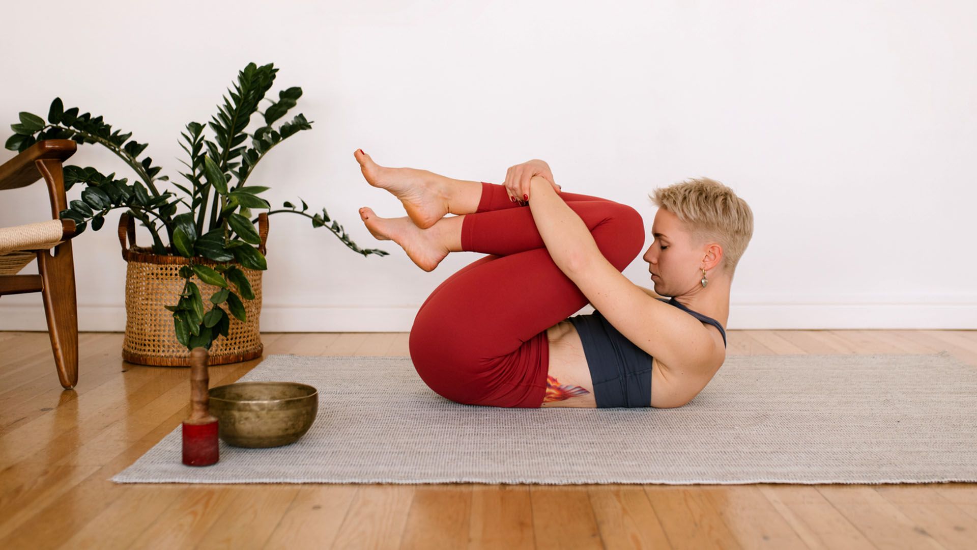 Woman with short blond hair and a tattoo on her side wearing red leggings and a black tank top does apanasana yoga pose, stretching her knees to her chest while lying down on a gray mat.