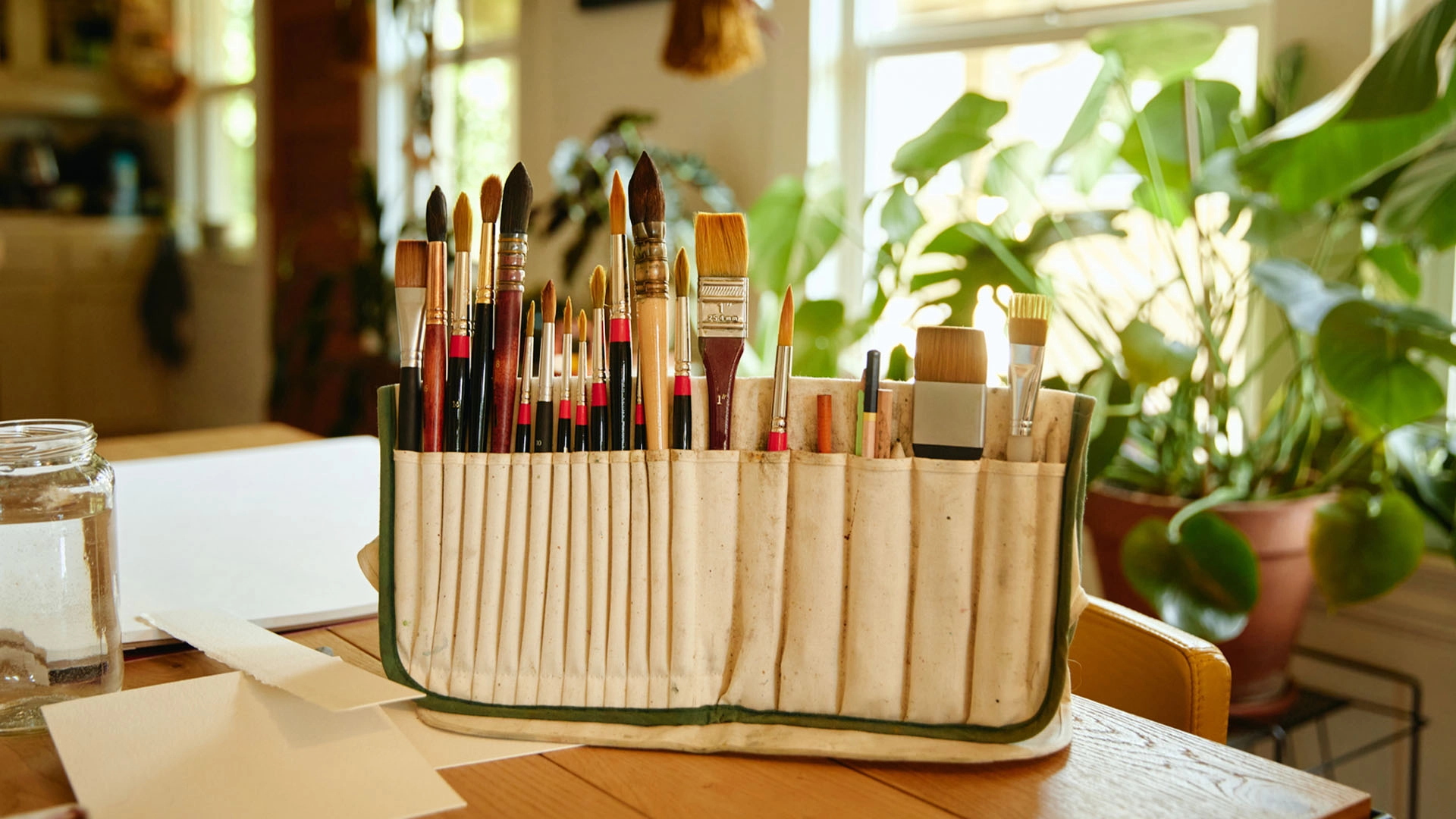 Line of paint brushes on a wooden table with plants and a sunny window in the background.