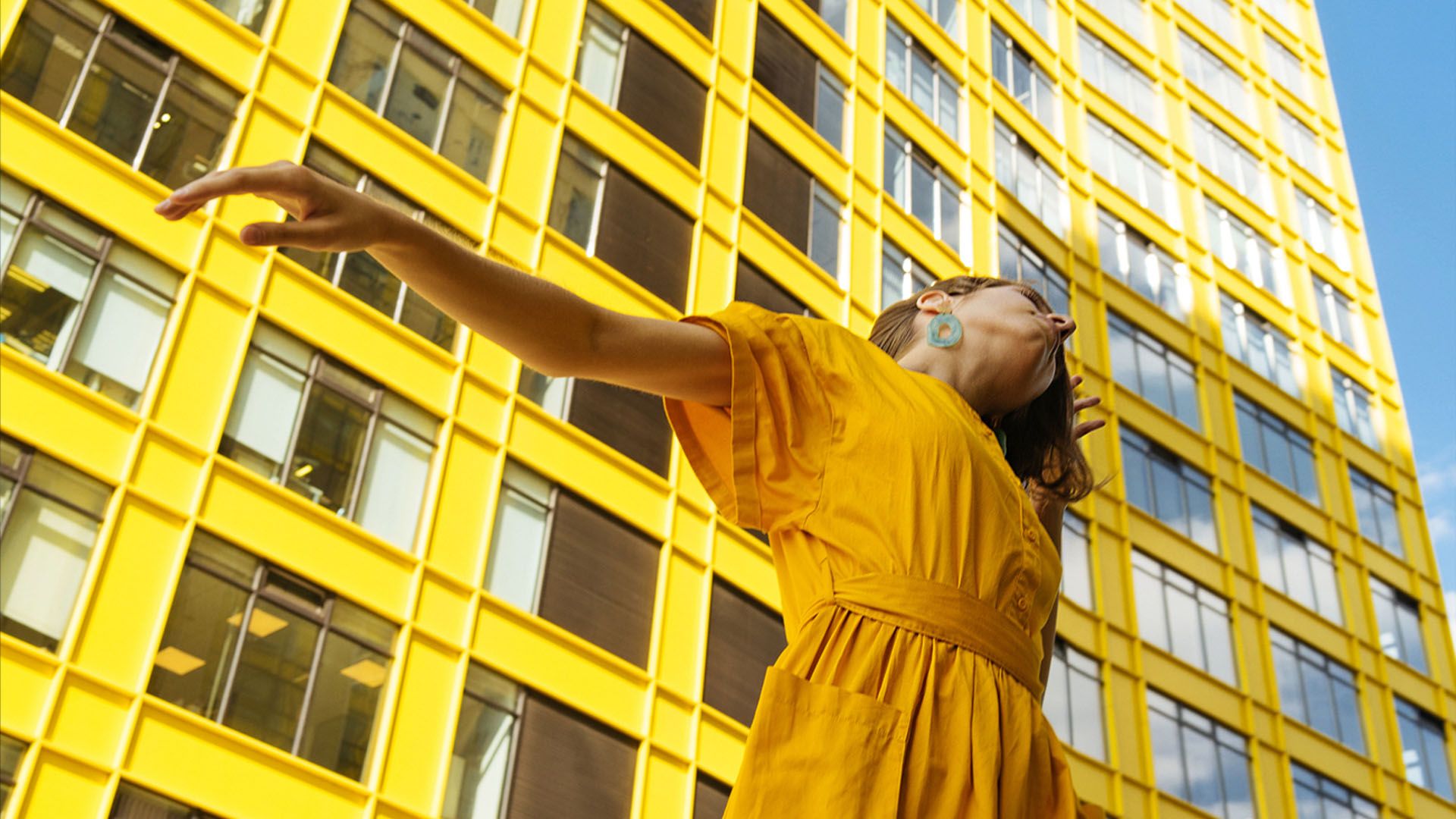 Woman in bright yellow dress and blue hoop earrings stands with one arm outstretched in front of a yellow skyscraper