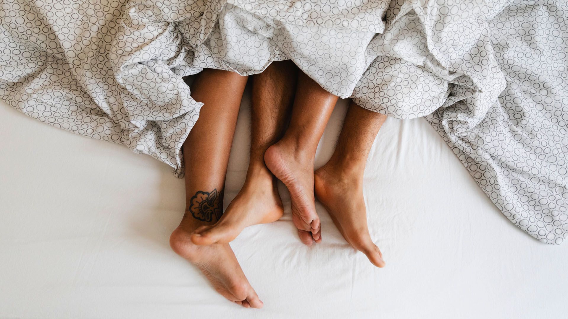 Detail of couple's feet closely intertwined shot from above as they lay in a bed with white and gray patterned sheets