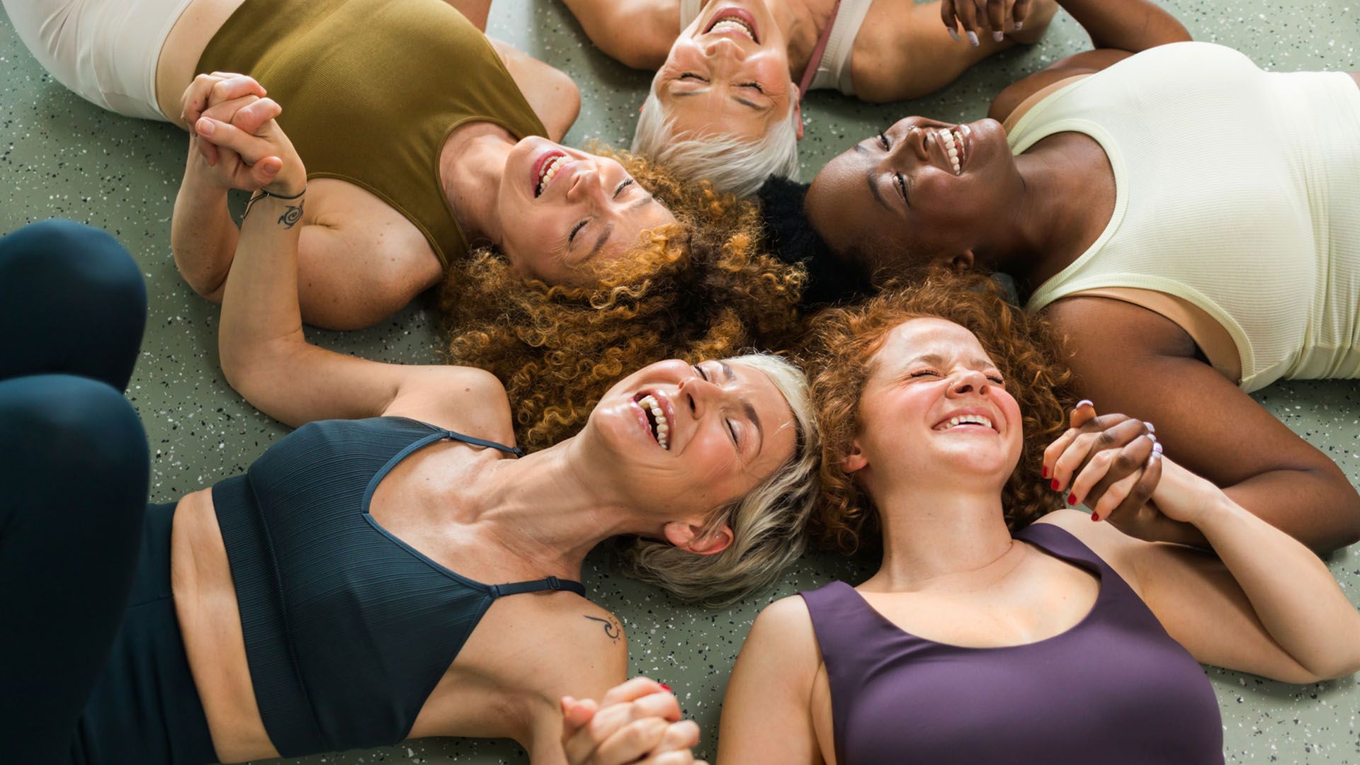 Top view of a circle of women of different ages and races smiling and holding hands while doing yoga and laughing.