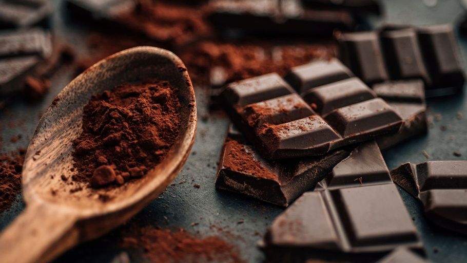 Close-up shot of squares of dark chocolate alongside a wooden spoon filled with cocoa powder.