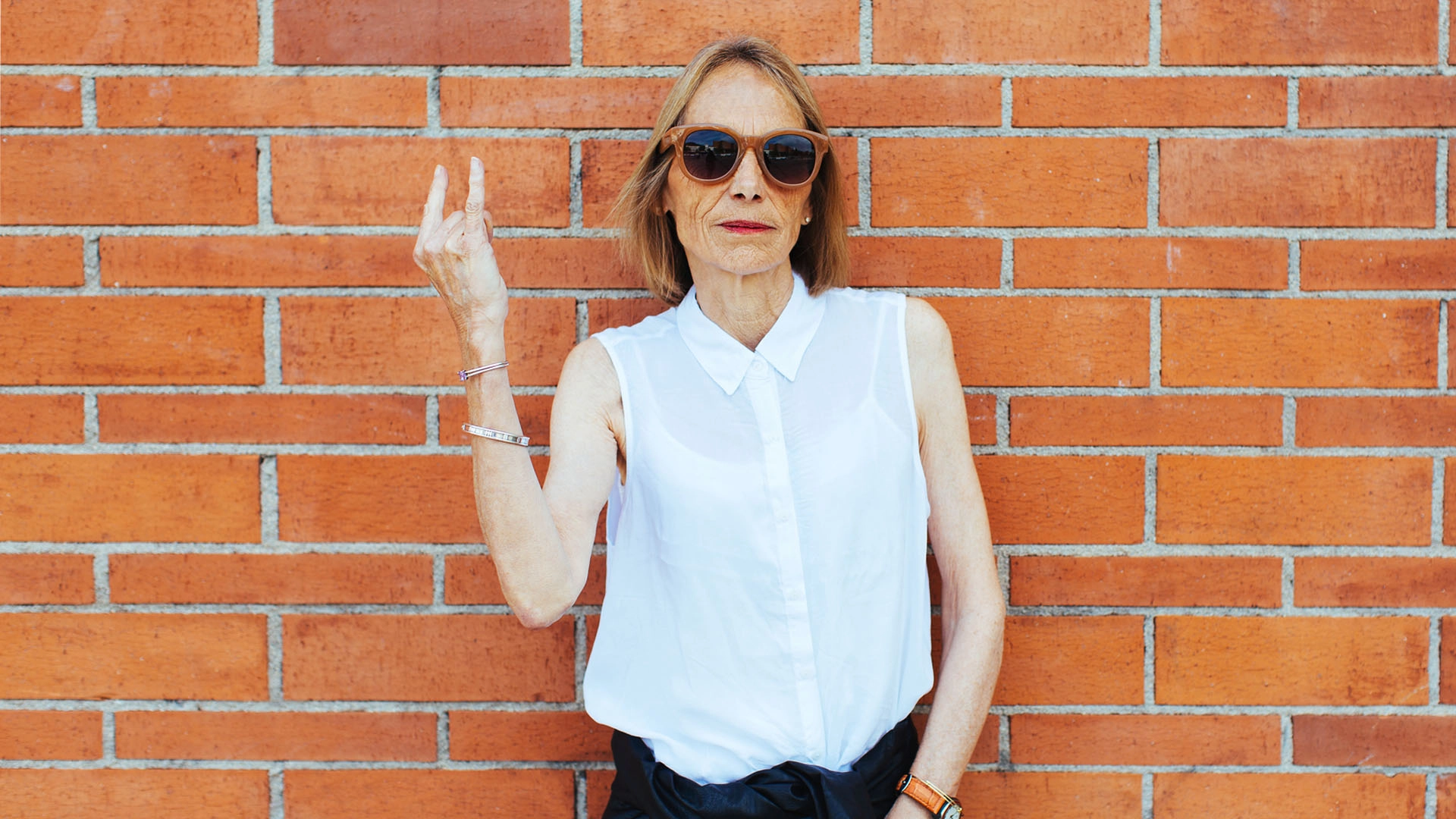 Senior woman with a serious expression wearing a sleeveless white button-down shirt and sunglasses stands against a brick wall, doing the rock ‘n’ roll sign with her right hand.