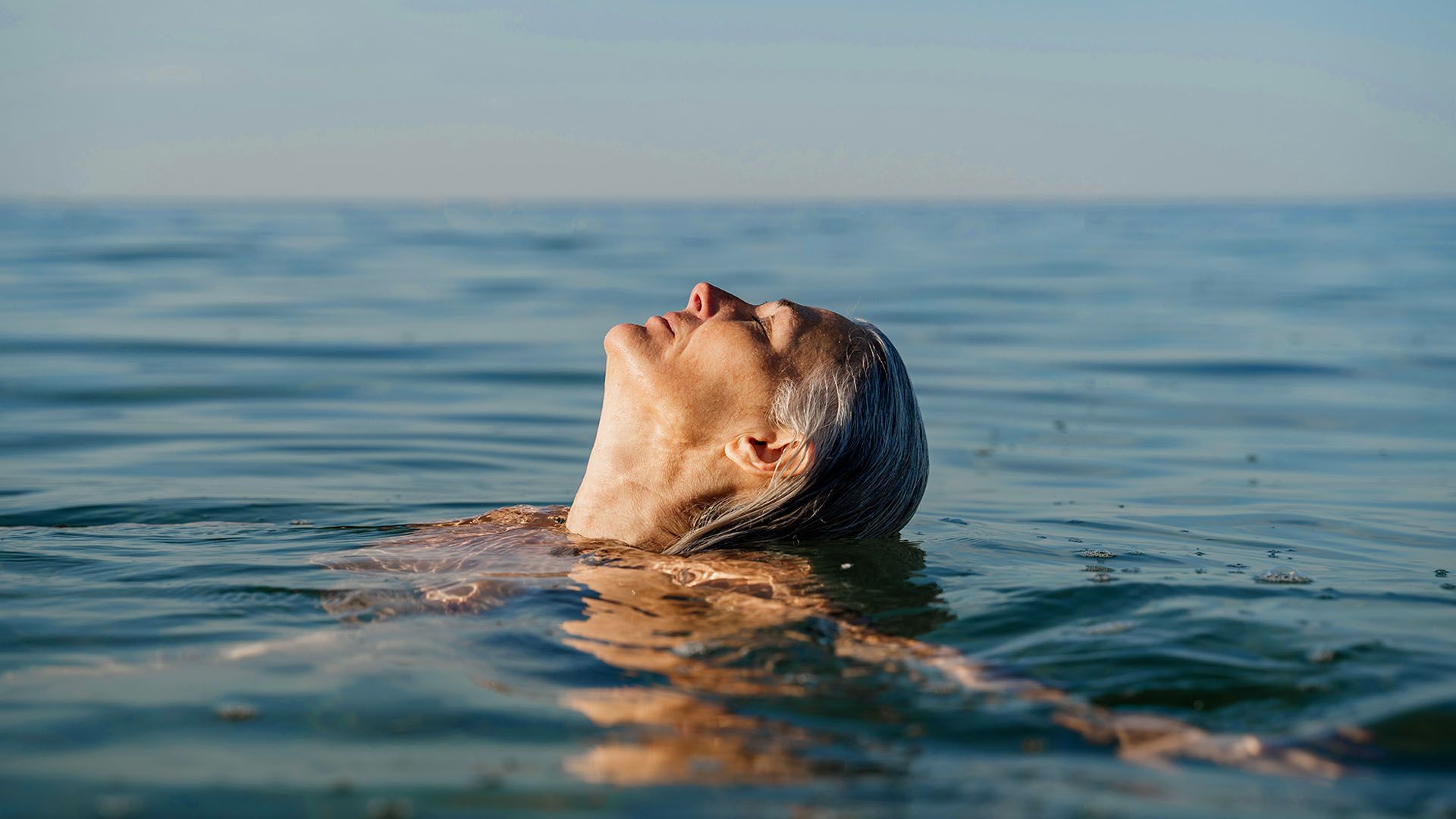 Mature woman floating and relaxing in open ocean, her head back and eyes closed, as she enjoys being in the water.