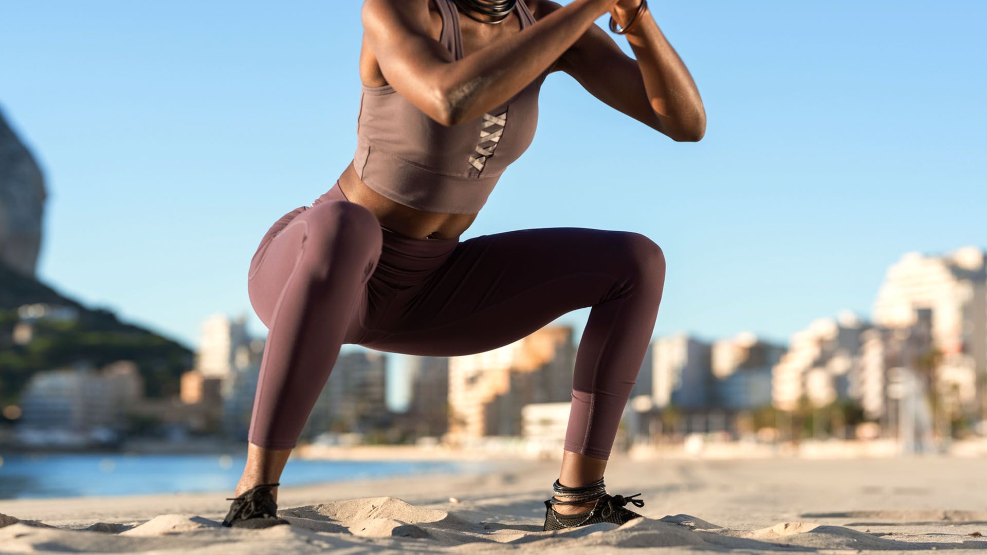 Female athlete wearing workout leggings and tank top does squats on a sandy beach with a bright blue sky above her.