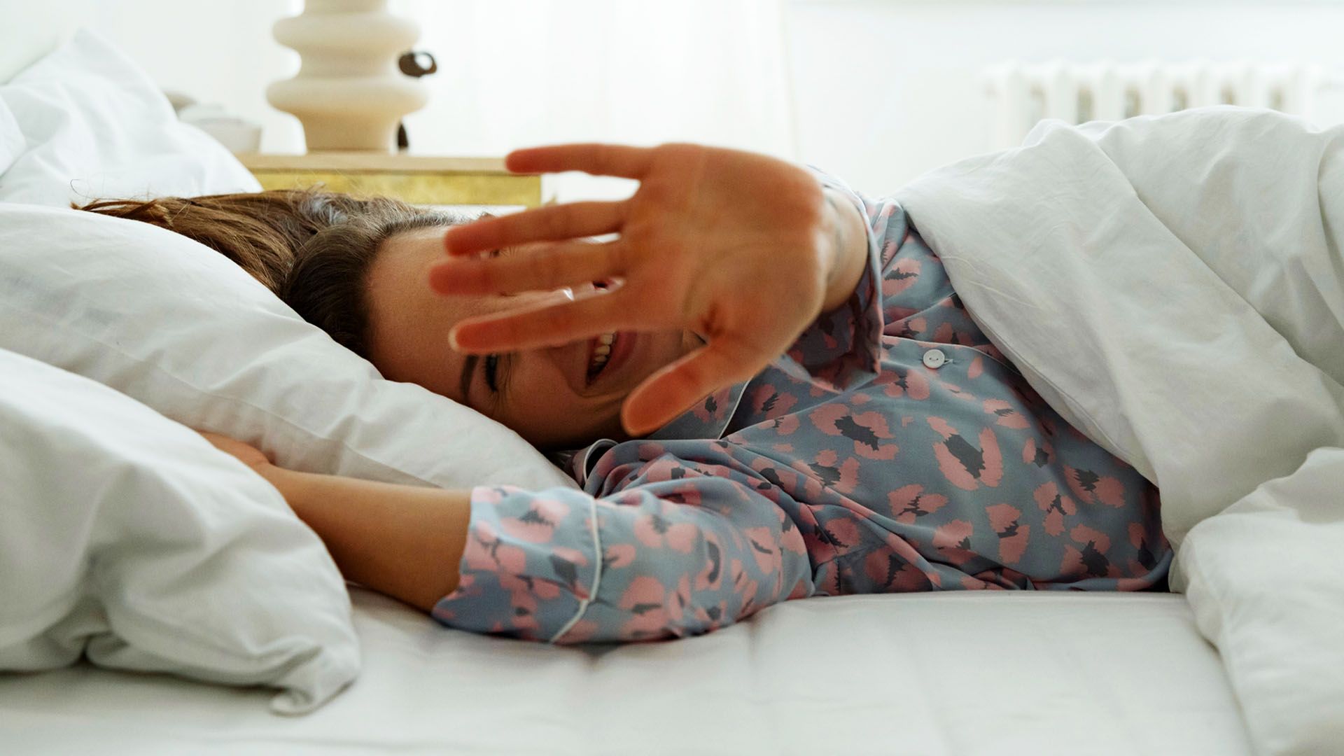 Postnatal woman wearing wedding ring lies in bed laughing, playfully holding her hand in front of her face