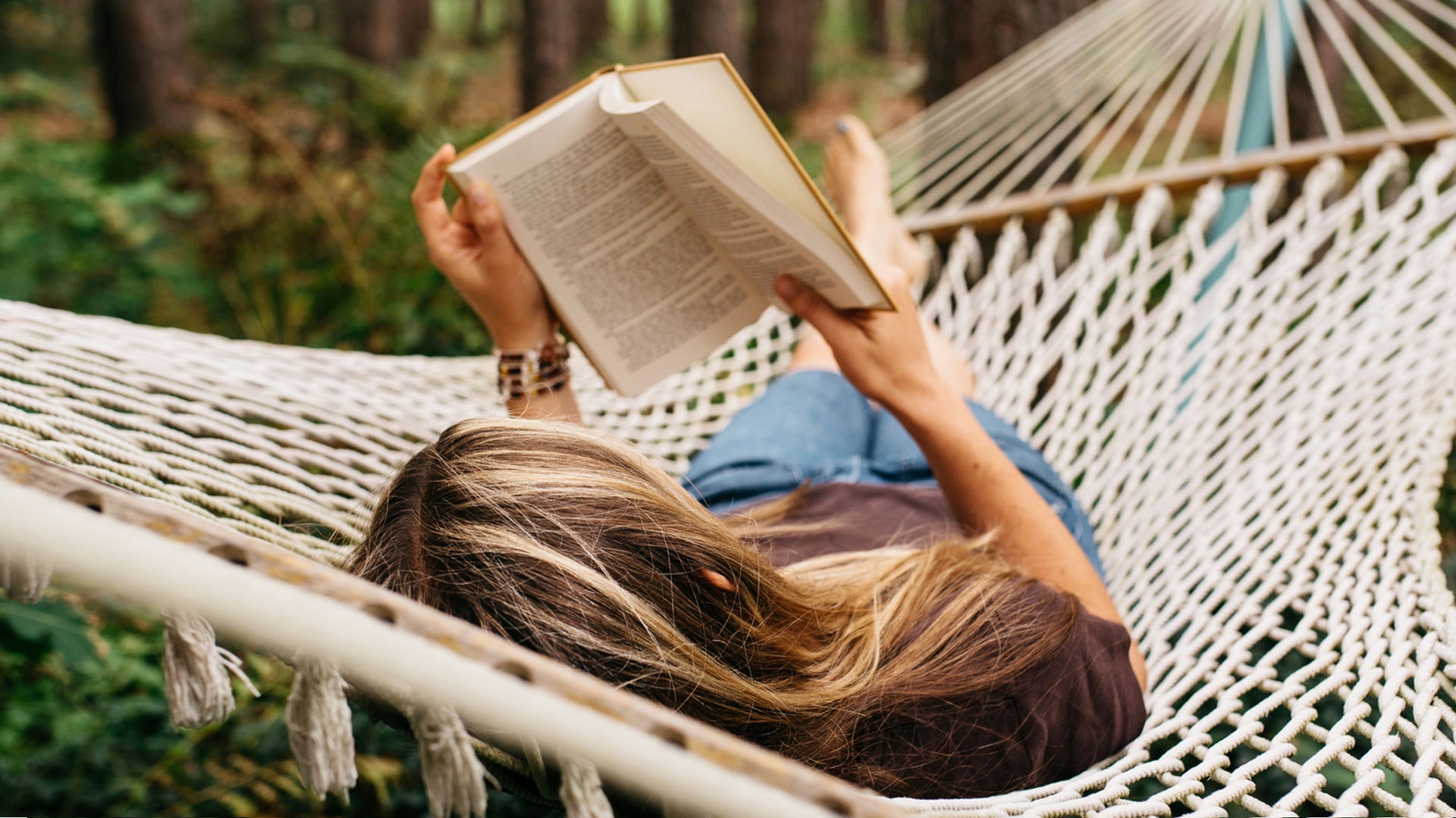 Woman lays in a white woven hammock outdoors, reading a book, practicing slow living rather than living a faster-paced lifestyle.