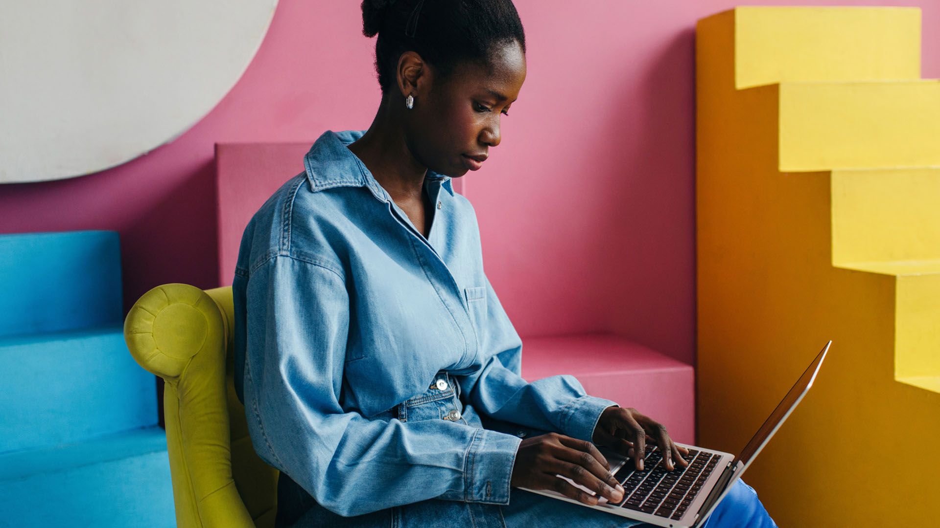 Black woman wearing blue shirt types on her laptop computer while seated in a colorful, modern space.