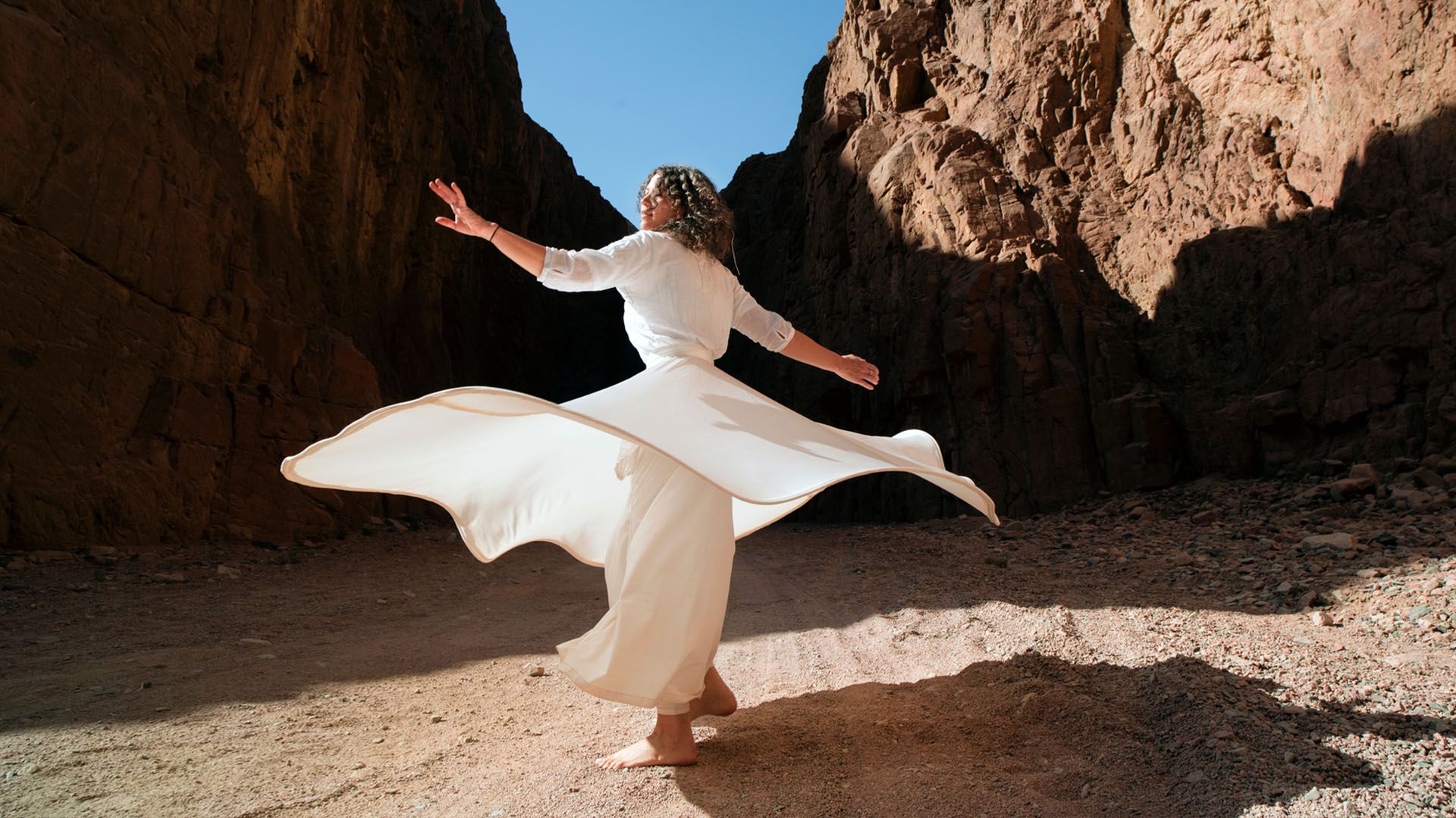 Woman in bare feet wearing a white dress that flows gently around her dances inside a canyon with a blue sky above her.