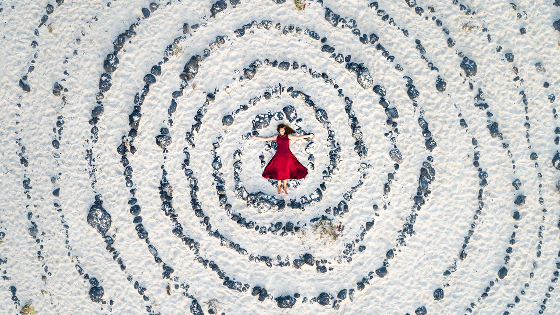 Woman in red dress contemplating the Venus retrograde surrounded by a spiral of rocks on sand.