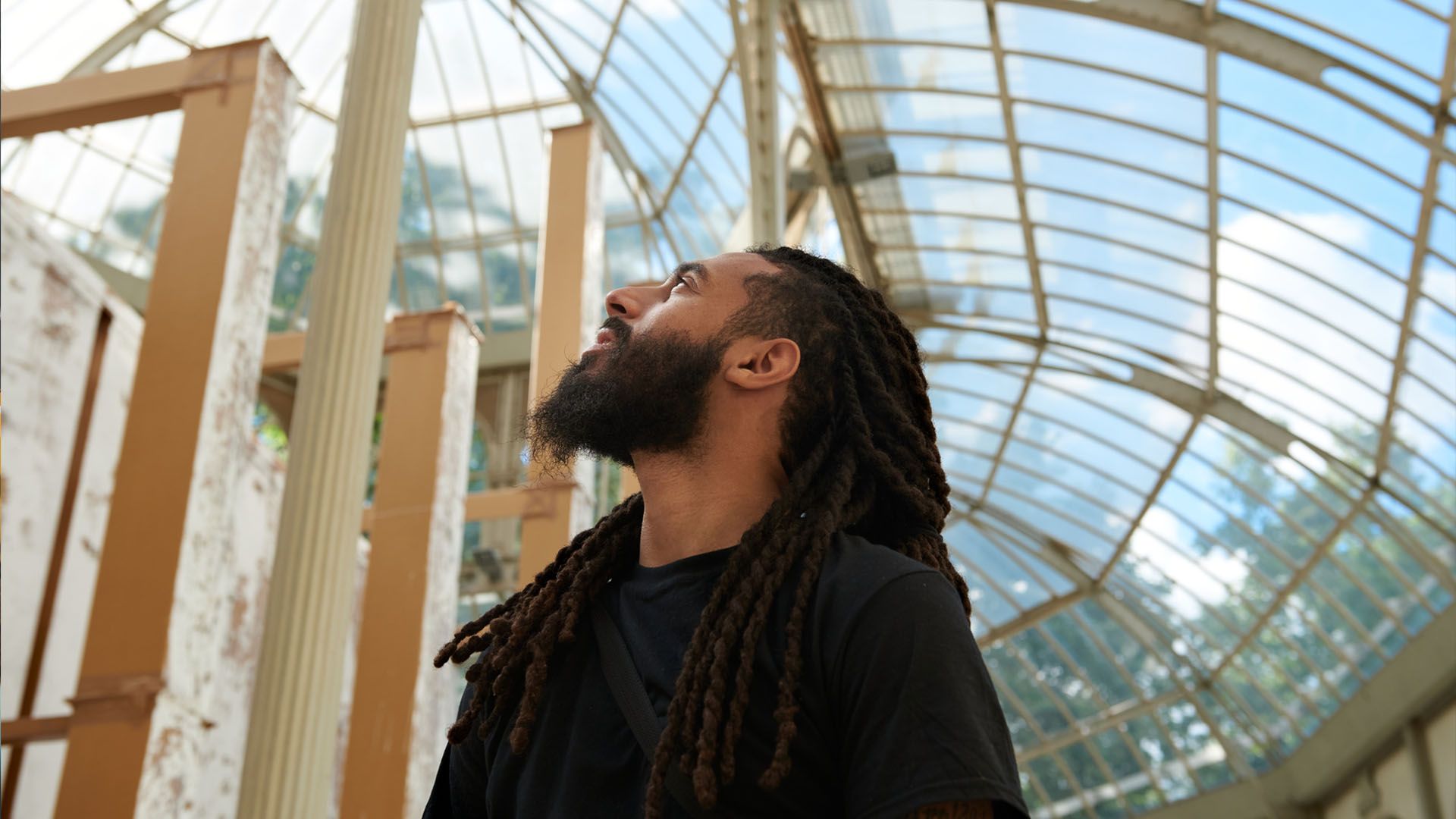 A Black man with a beard and dreadlocks wearing a short-sleeved t-shirt stands underneath a glass-domed ceiling and looks with curiosity towards the sky on a sunny day.